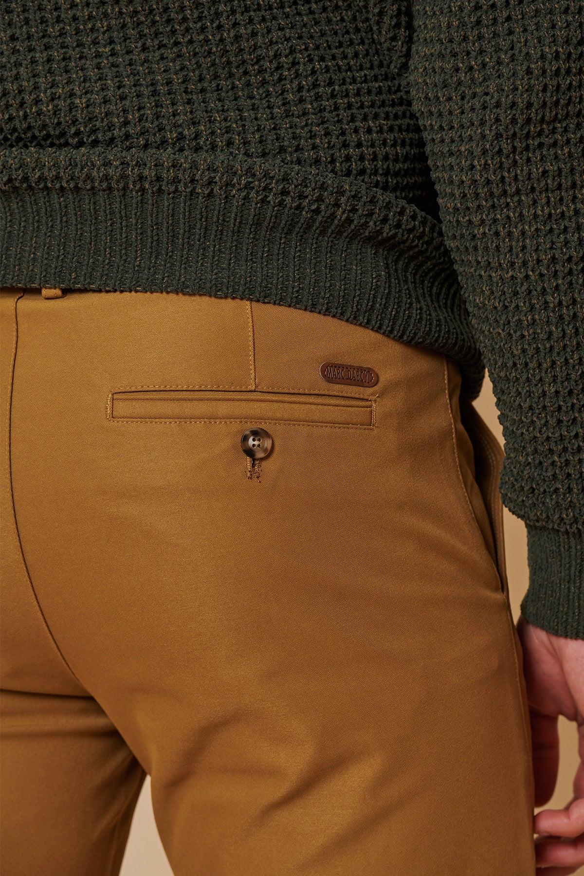 Close-up of a person wearing marcdarcy ENZO Tan Chinos with a buttoned back pocket and small brown label, paired with a textured dark green sweater. Only part of the person's back and right arm are visible.