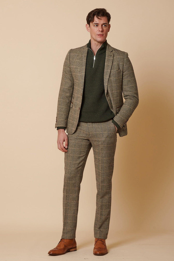 A man with short brown hair stands against a beige background in the Marc Darcy TED Tan Tweed Check Two Piece Suit, paired with a green zip-up sweater and brown leather dress shoes. He has a relaxed posture, one hand in his pocket.