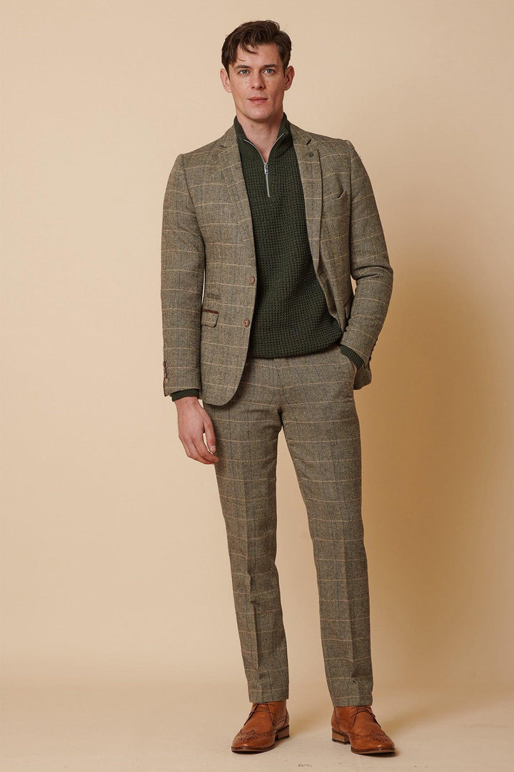 A man stands against a beige background wearing the Marc Darcy TED Tan Tweed Check Two Piece Suit, styled with a green quarter-zip sweater and brown dress shoes, one hand in his pocket.