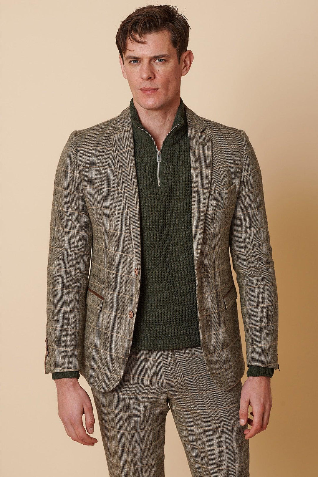 A man in the Marc Darcy TED Tan Tweed Check Two Piece Suit stands against a beige background, wearing a green knit zip-up sweater underneath and channeling a subtle Peaky Blinders style with a neutral expression.