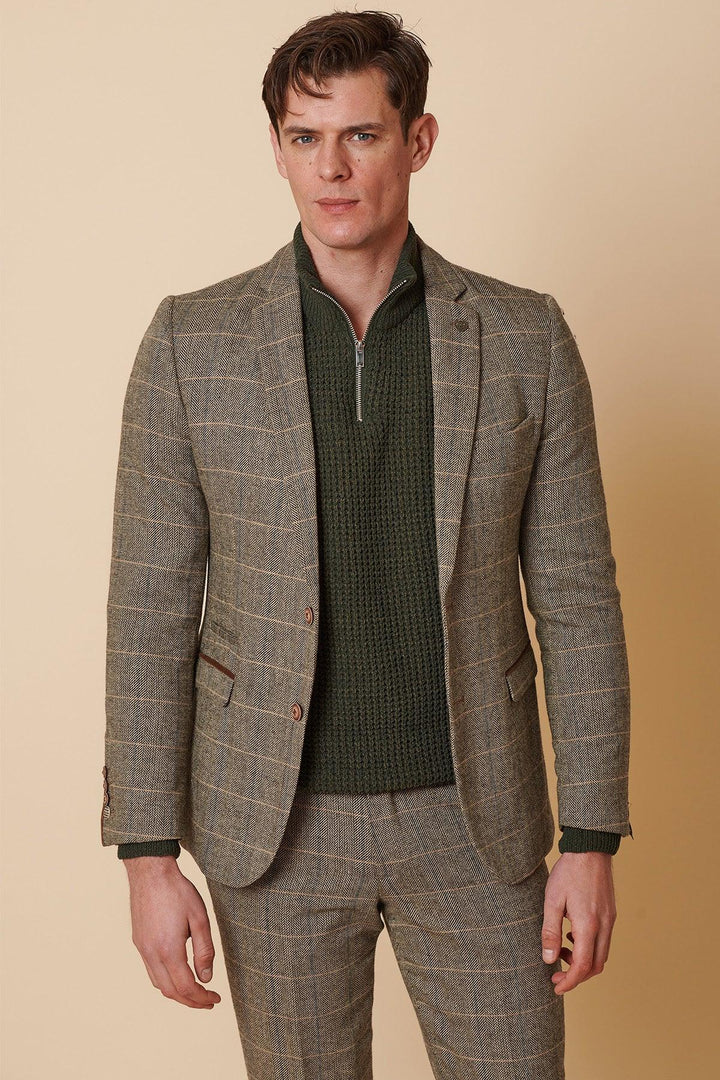 A man in the Marc Darcy TED Tan Tweed Check Two Piece Suit stands against a beige background, wearing a green knit zip-up sweater underneath and channeling a subtle Peaky Blinders style with a neutral expression.