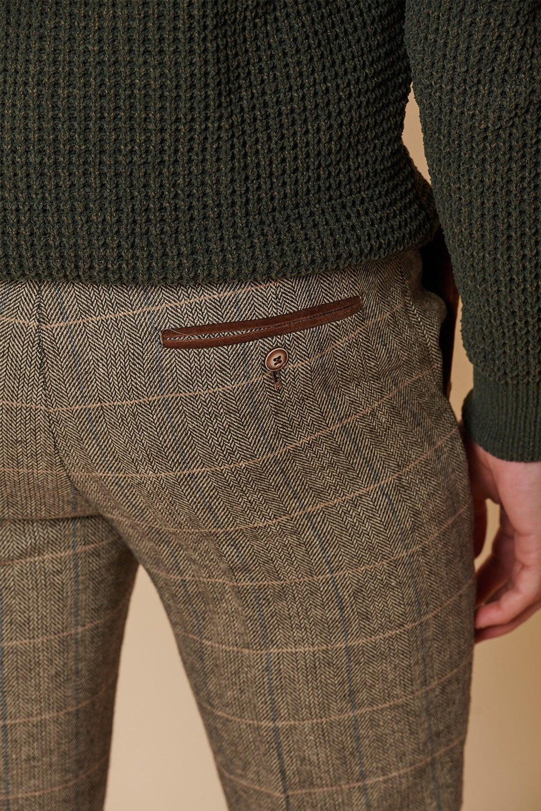 A person wearing marcdarcy's DX7 Tan Tweed Check Two Piece Suit, with brown plaid trousers featuring a leather-accented back pocket and button, paired with a dark green textured sweater—a perfect herringbone slim fit look.