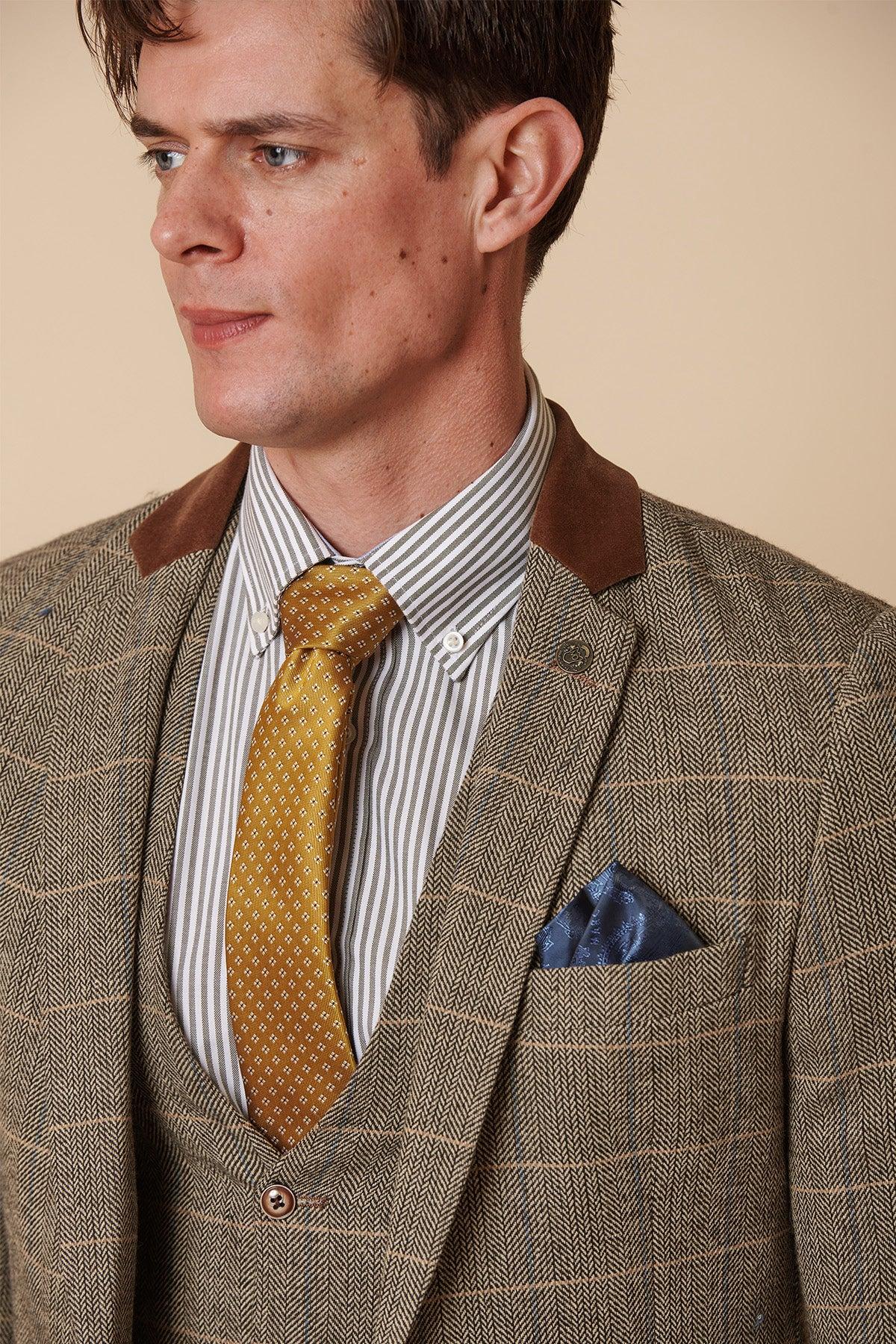 A man models the marcdarcy DX7 - Tan Tweed Check Two Piece Suit, styled with a striped shirt, yellow patterned tie, and blue pocket square, against a beige background.