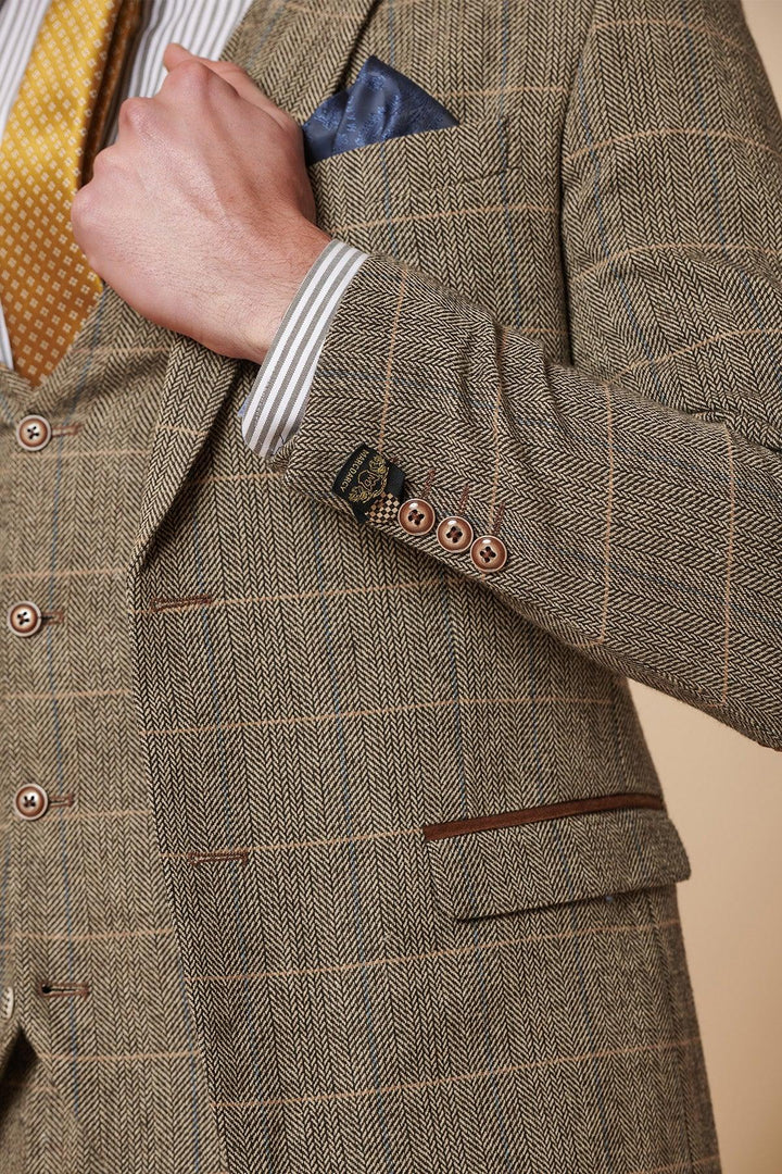 A close-up of a person in the Marc Darcy TED Tan Tweed Check Two Piece Suit, styled with a yellow patterned tie, striped shirt, and blue pocket square, adjusting their lapel for a classic Peaky Blinders-inspired look.