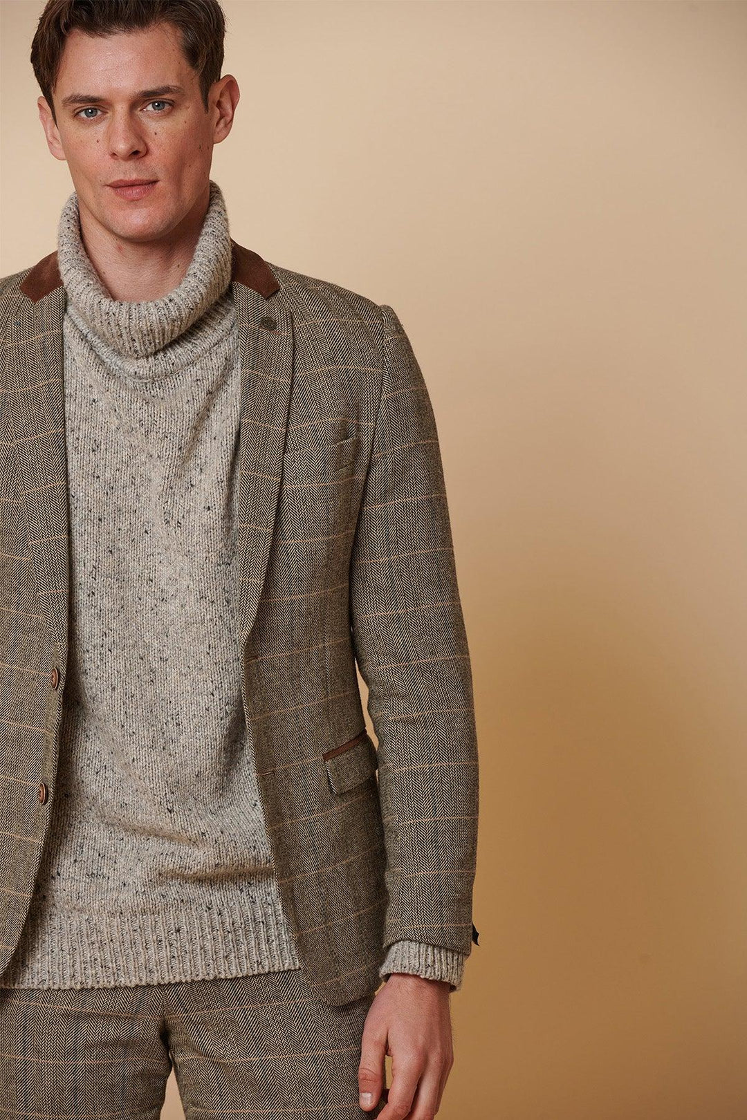 A man in the marcdarcy DX7 - Tan Tweed Check Two Piece Suit stands against a beige background, facing the camera with a neutral expression, wearing brown accents and a chunky light gray turtleneck sweater.