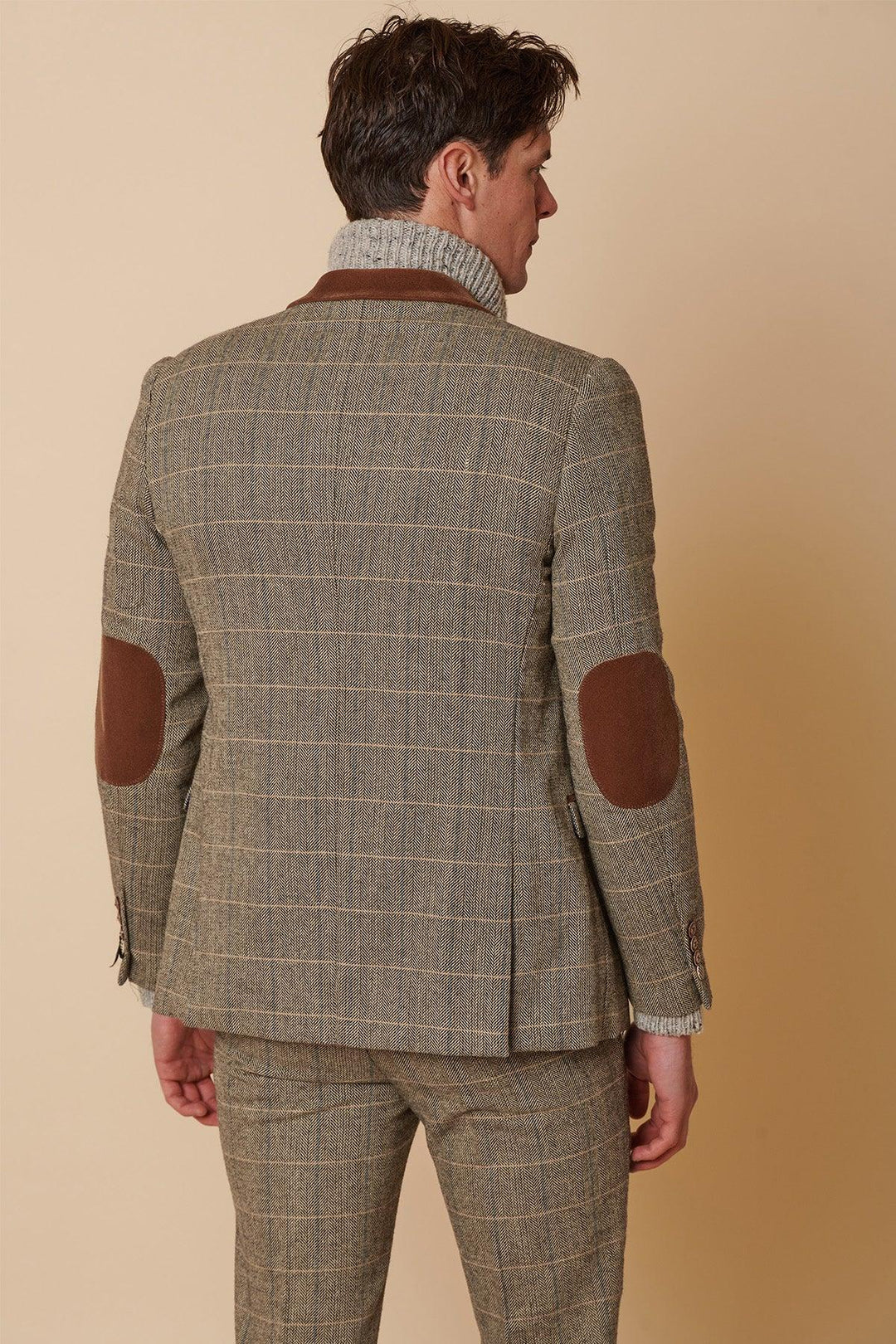 A man in the marcdarcy DX7 - Tan Tweed Check Two Piece Suit stands with his back to the camera, wearing matching trousers, brown elbow patches on the blazer, and a light grey turtleneck underneath.