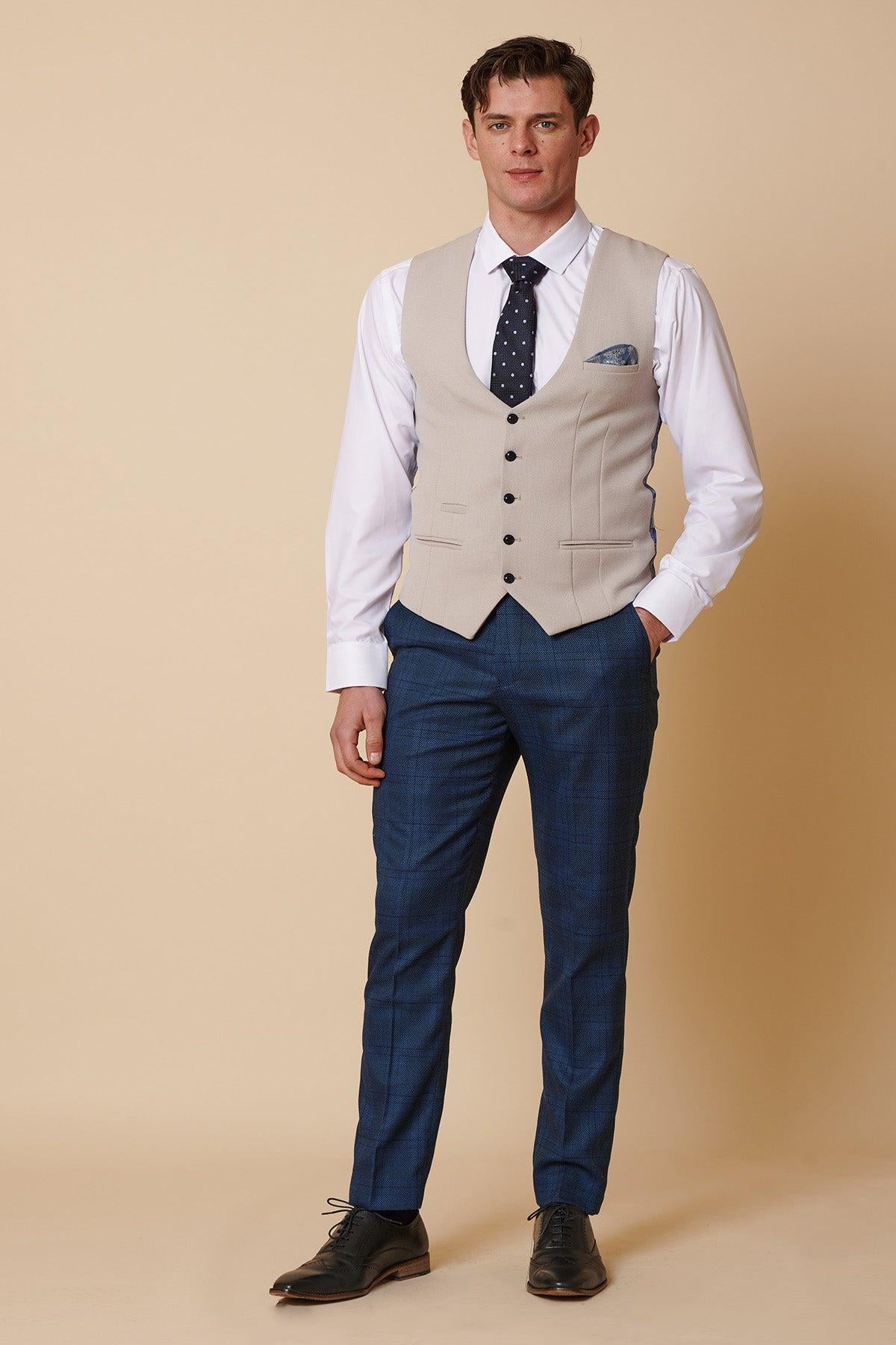 A man stands against a beige background wearing the marcdarcy KELVIN Stone Single Breasted Waistcoat, a white dress shirt, navy blue patterned tie, blue plaid trousers, pocket square, and black dress shoes. He poses smartly with one hand in his pocket.
