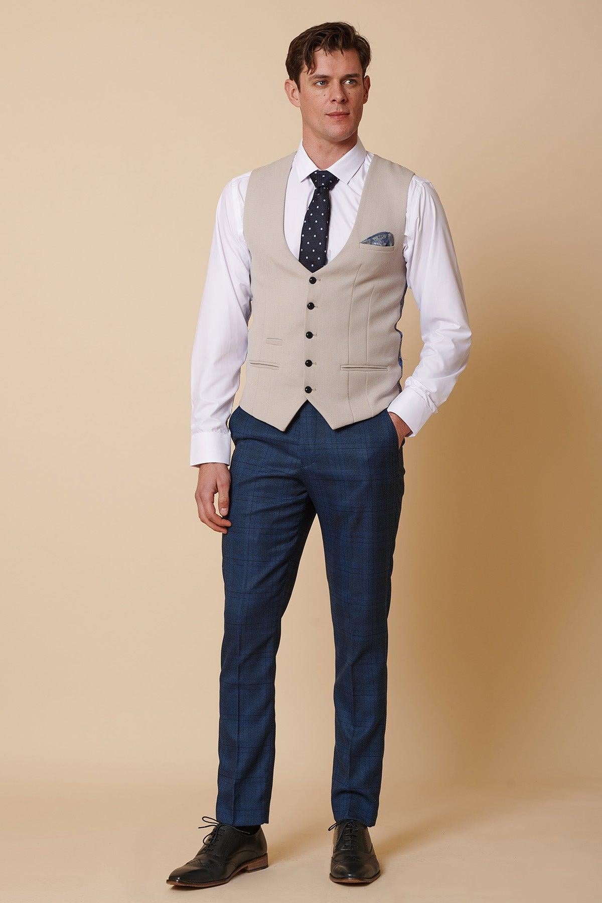 A man stands against a beige background wearing the marcdarcy KELVIN Stone Single Breasted Waistcoat over a white shirt, paired with a navy polka dot tie, blue plaid trousers, black dress shoes, and a pocket square for refined formalwear.