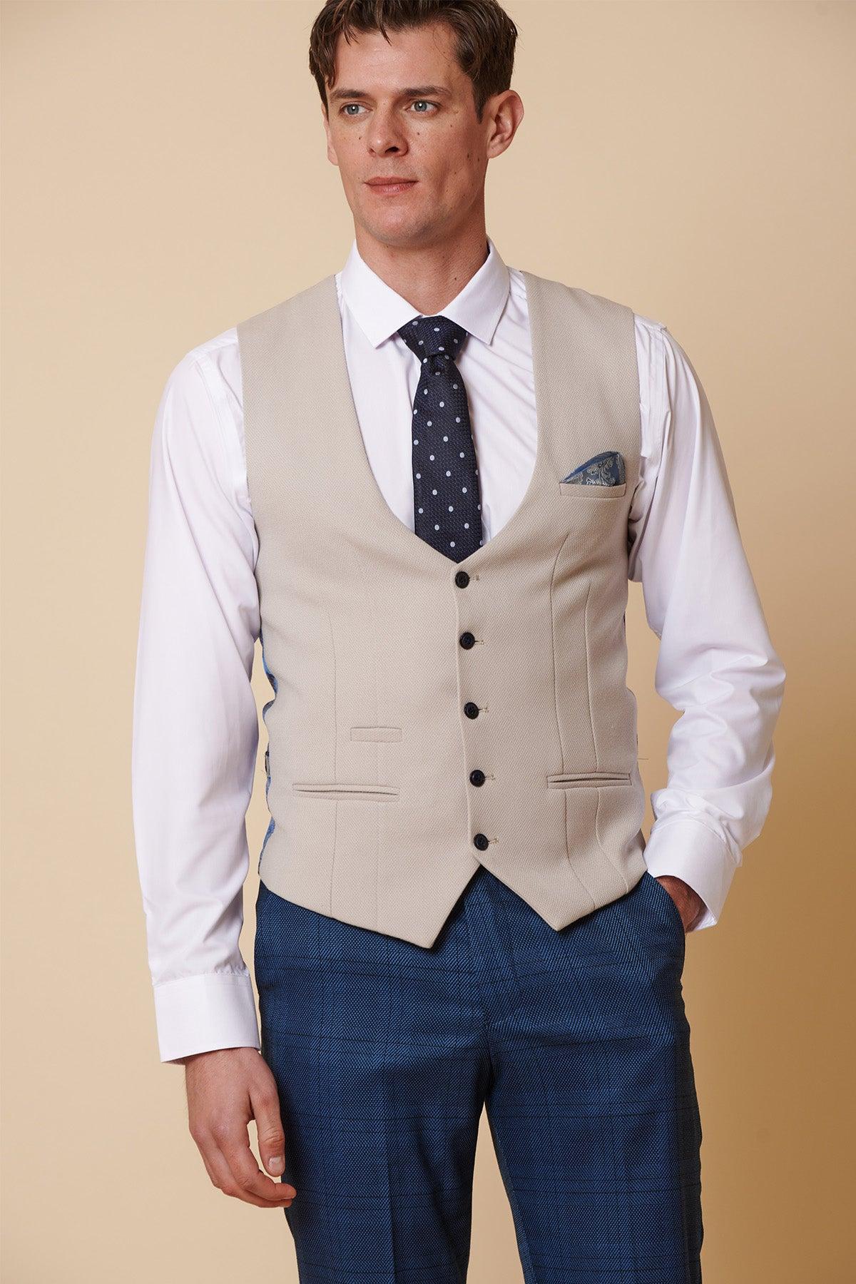 A man models the marcdarcy KELVIN - Stone Single Breasted Waistcoat over a white shirt, navy polka dot tie, pocket square, and blue plaid dress pants against a beige background, highlighting elegant formalwear.