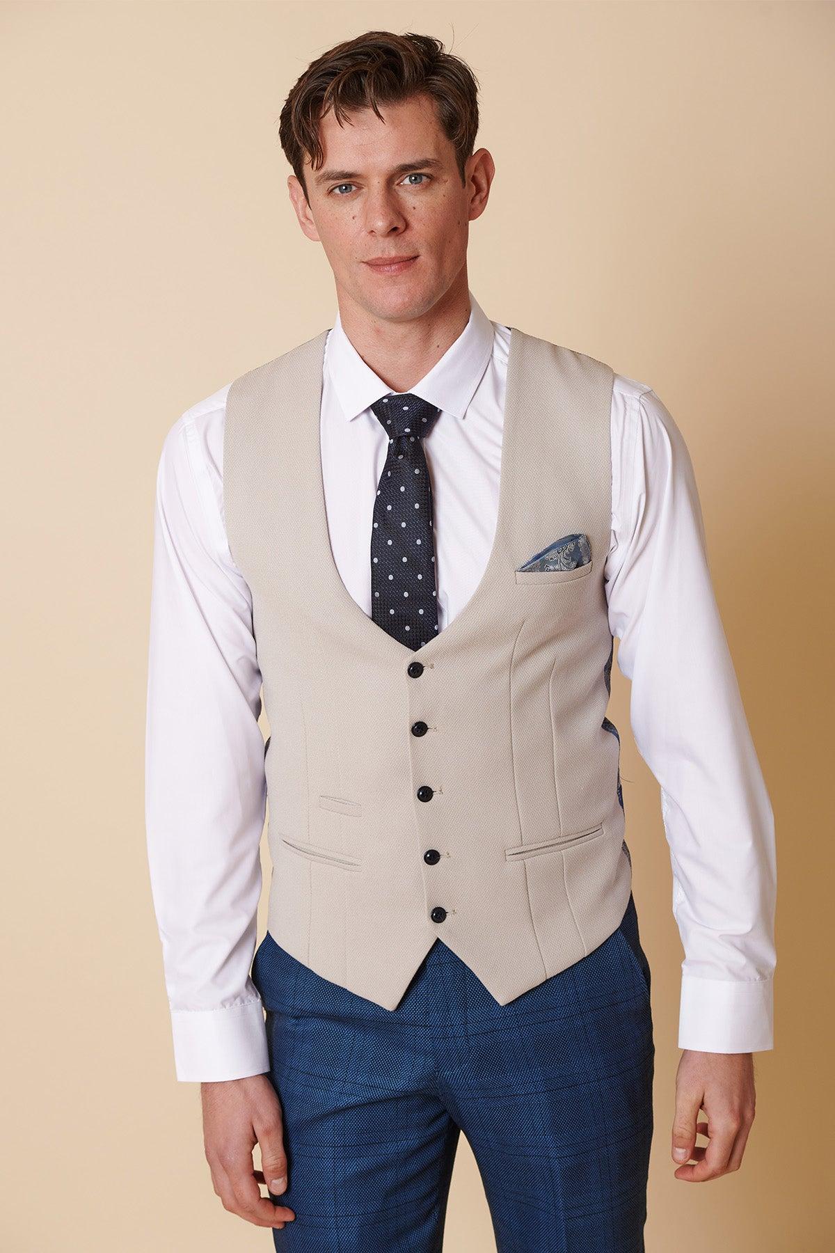 A man models the marcdarcy KELVIN - Stone Single Breasted Waistcoat over a white shirt with a navy polka dot tie, blue plaid trousers, and pocket square, posing against a beige background to showcase refined formal style.