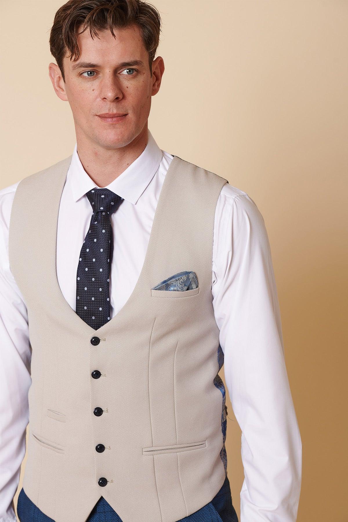 A man wears the marcdarcy KELVIN Stone Single Breasted Waistcoat over a white shirt, paired with a navy polka dot tie and patterned pocket square, standing against a plain light tan background.
