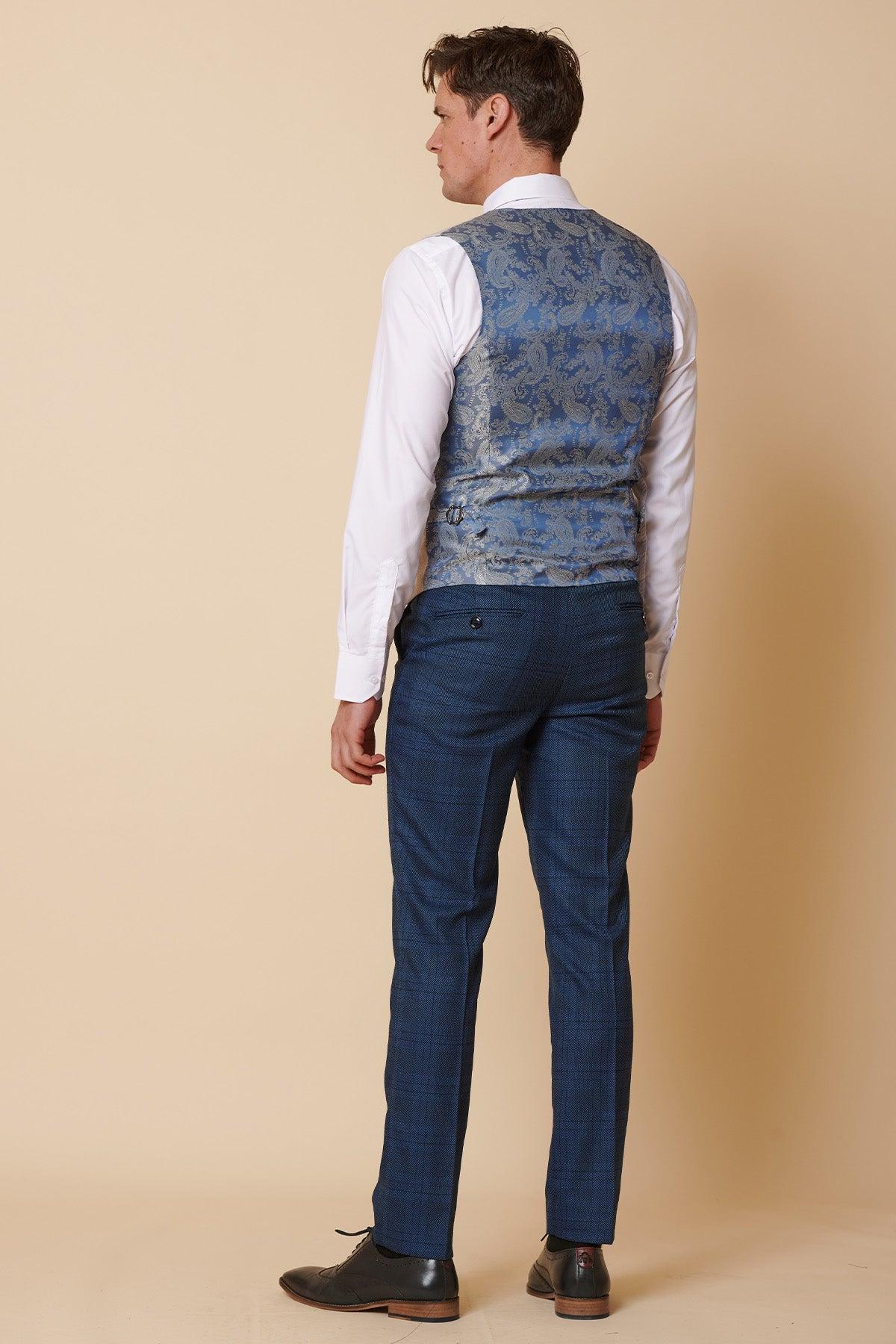 A man stands facing away in slim fit formalwear, wearing a white shirt, marcdarcy KELVIN Stone Single Breasted Waistcoat, blue plaid trousers, and black shoes against a beige background.
