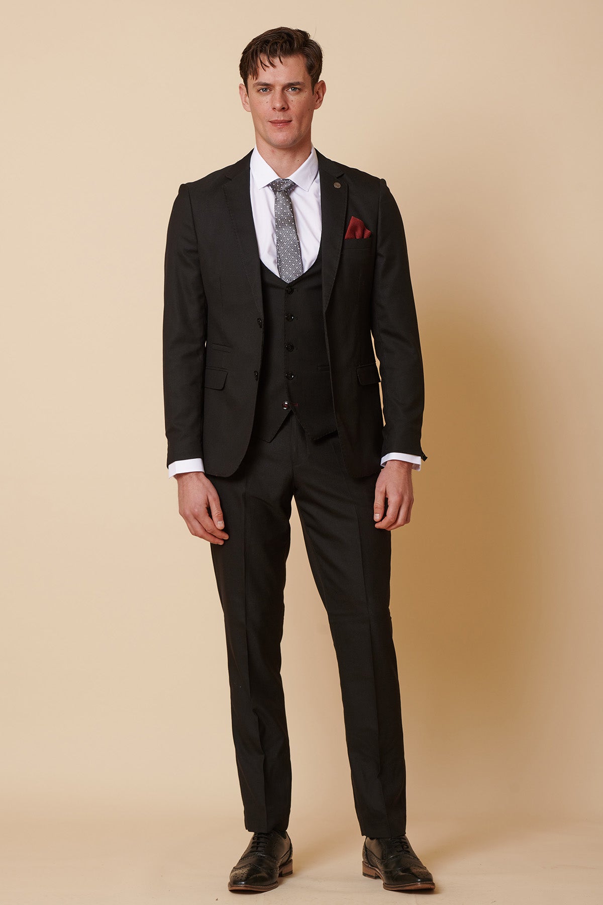 A man faces forward against a beige background wearing the Marc Darcy DANNY black three-piece suit with single-breasted waistcoat, white shirt, gray patterned tie, and red pocket square, showcasing timeless formal style.