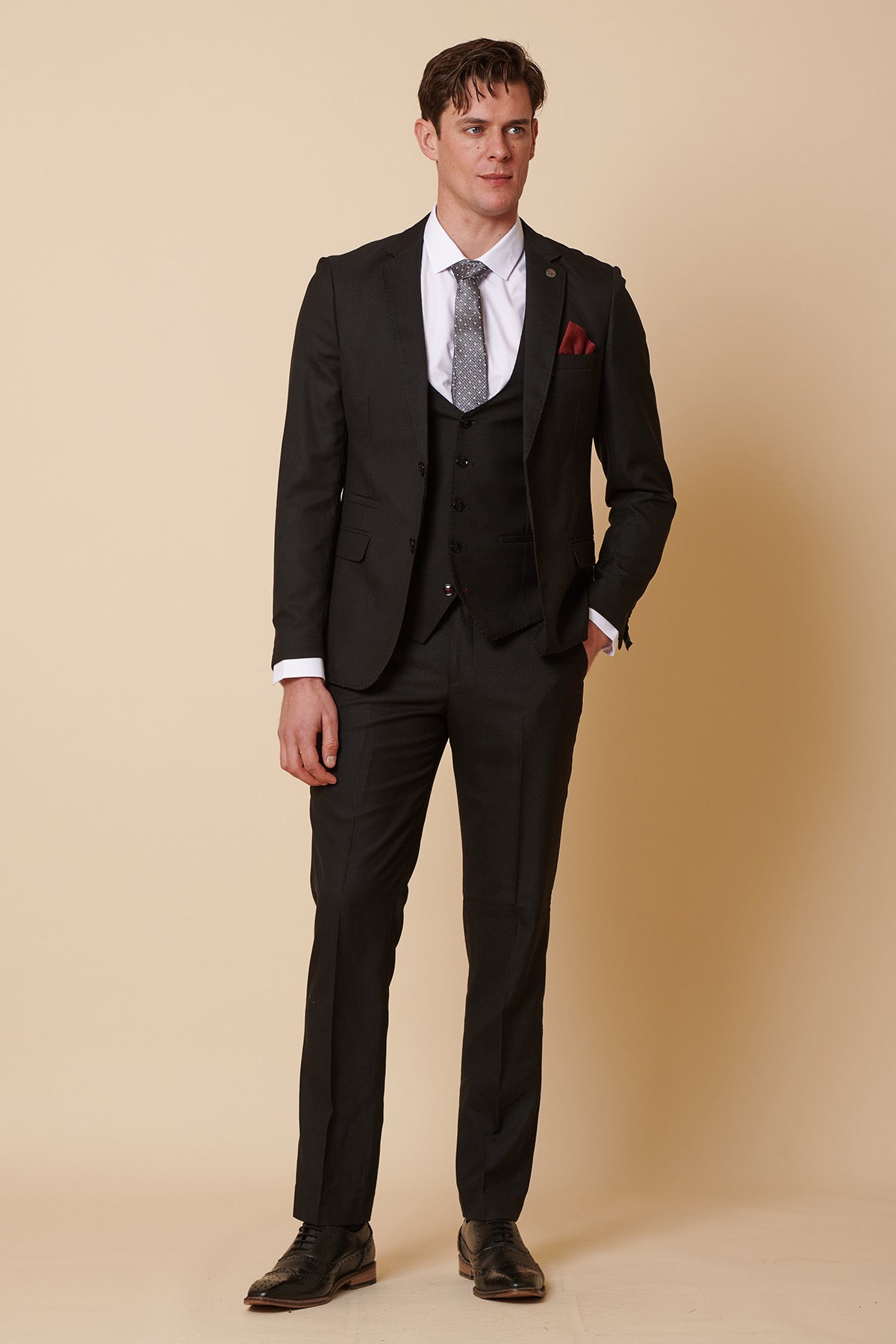 A man stands against a beige background wearing the Marc Darcy DANNY black three-piece suit with single breasted waistcoat, white shirt, gray patterned tie, red pocket square, and black dress shoes—a classic formal look.