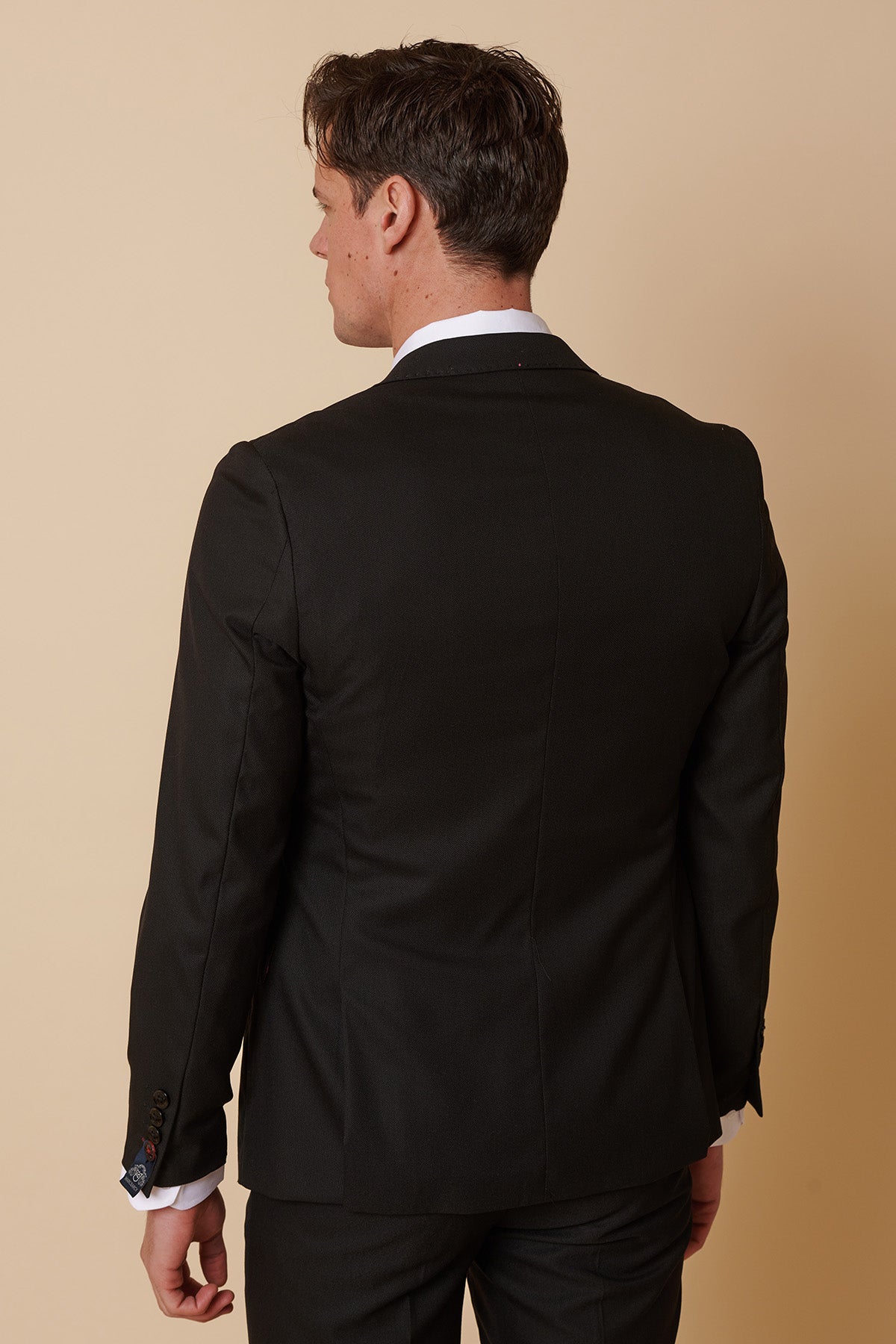 A man in the Marc Darcy DANNY Black Three Piece Suit with a single breasted waistcoat and white shirt stands facing away, highlighting the back of his tailored outfit against a beige background.