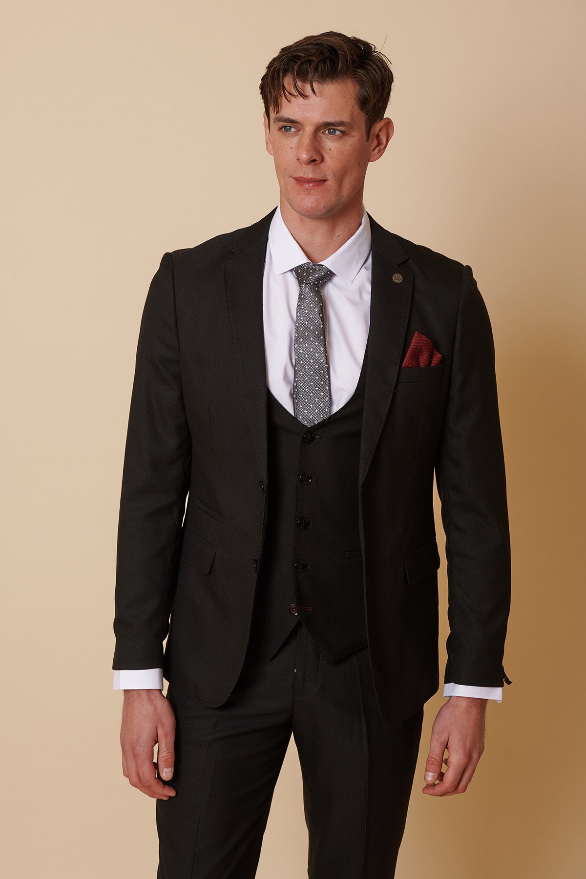 A man stands against a beige background wearing the Marc Darcy DANNY black three piece suit with single breasted waistcoat, a white dress shirt, patterned gray tie, and red pocket square—a sharp choice for any formal occasion.