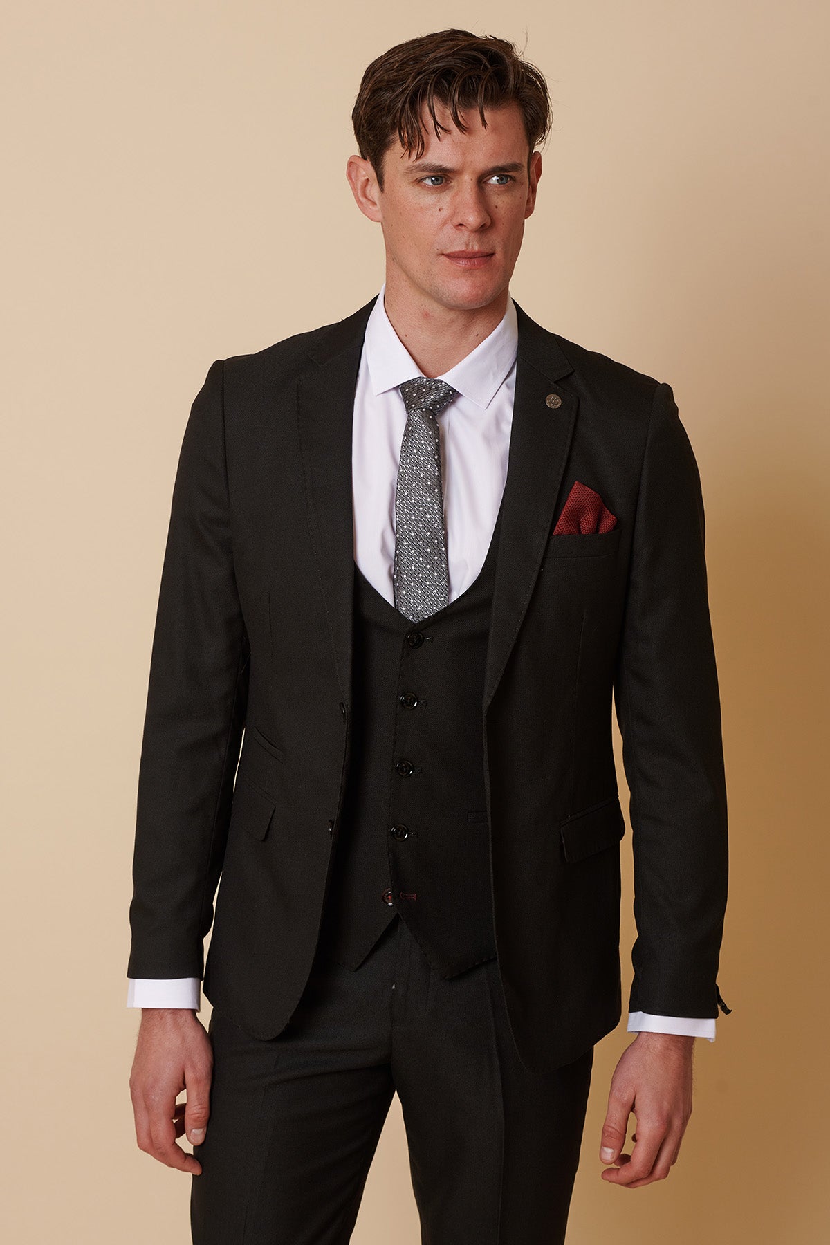 A man wearing a Marc Darcy DANNY Black Tailored Blazer with a matching vest, white dress shirt, patterned gray tie, and red pocket square stands against a beige background.