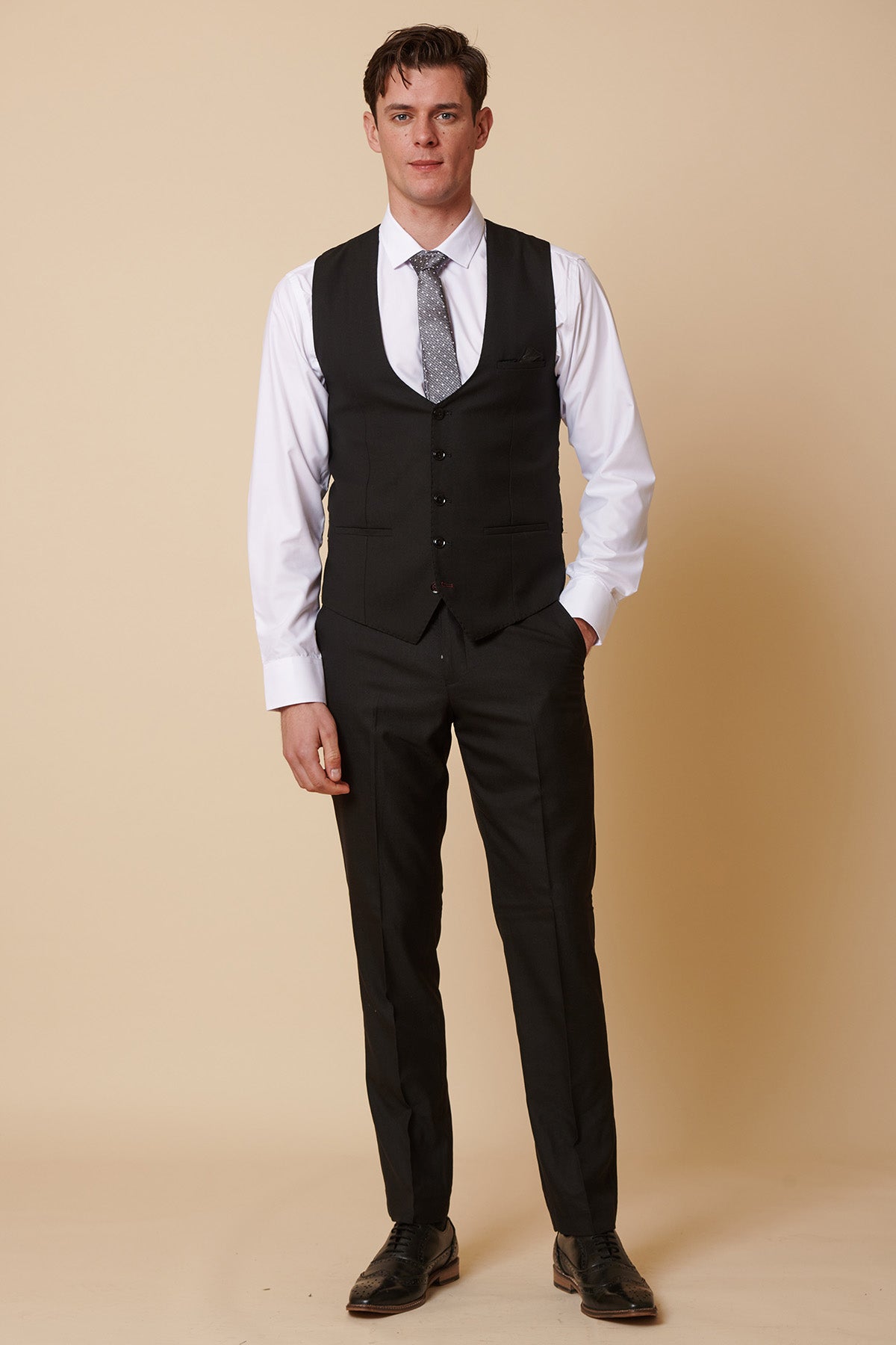 A man poses against a beige background in the Marc Darcy DANNY black three-piece suit with a single-breasted waistcoat, paired with a white shirt, silver tie, and black shoes—capturing timeless formal style.