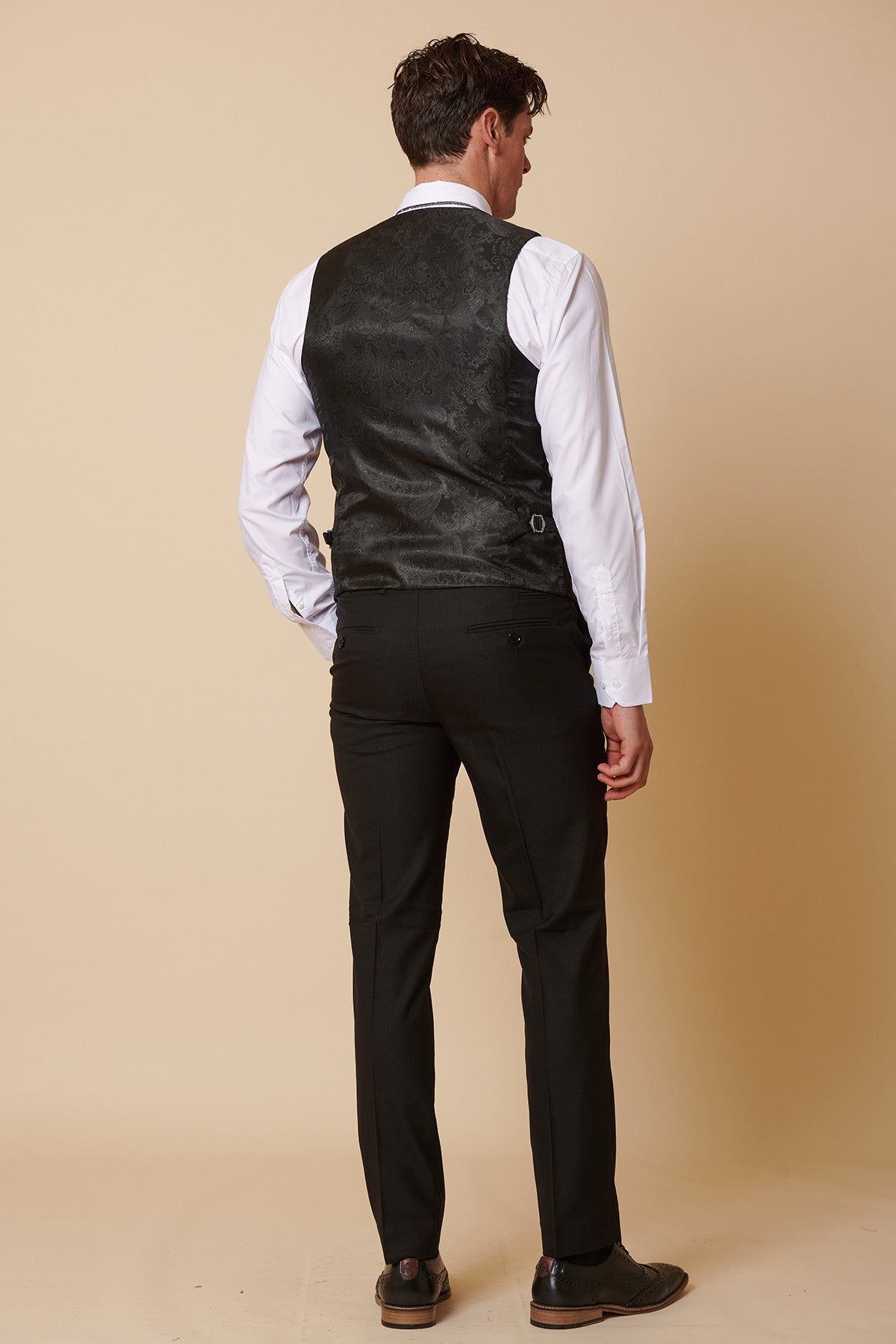 A man, dressed for a formal occasion in the Marc Darcy DANNY Black Three Piece Suit with Single Breasted Waistcoat, stands facing away from the camera against a plain beige background.