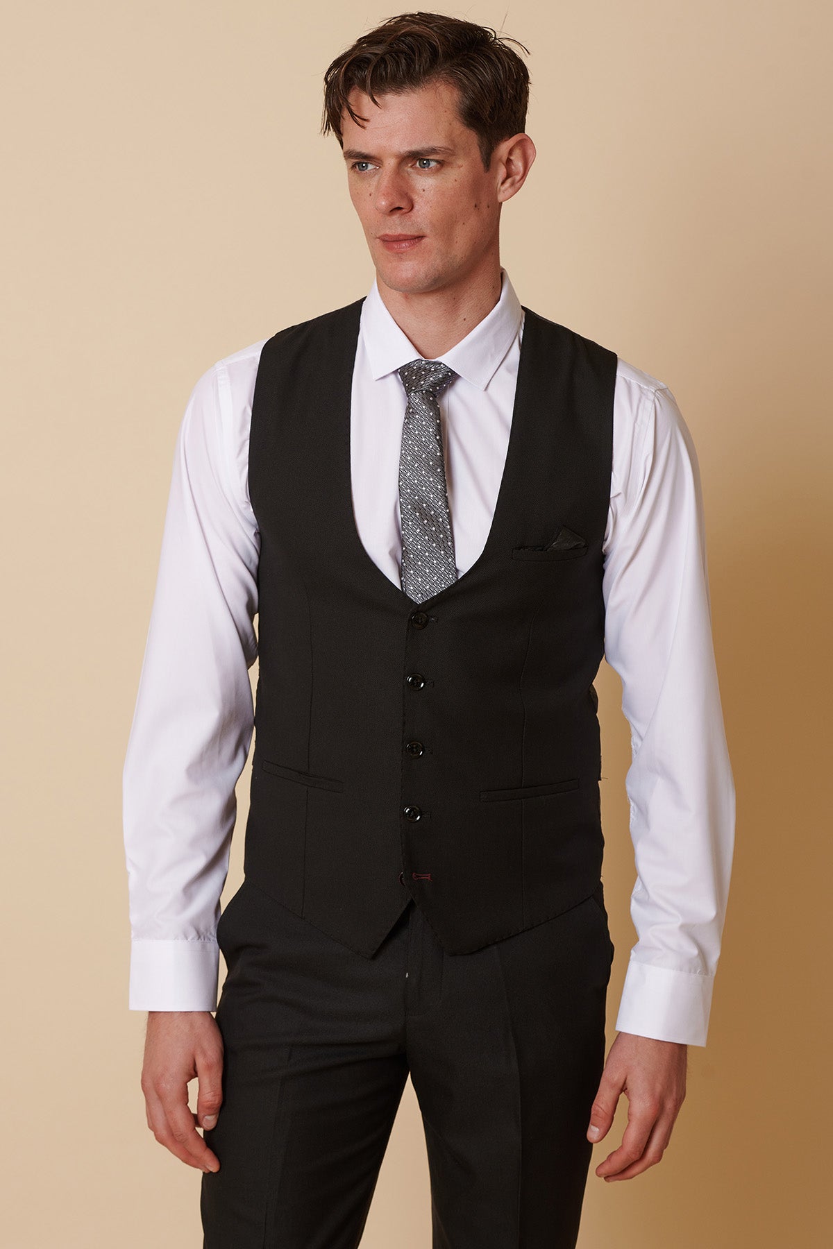 A man stands against a beige background, wearing the Marc Darcy DANNY black three piece suit with a single breasted waistcoat, white shirt, patterned gray tie, and black dress pants, looking slightly to the side.