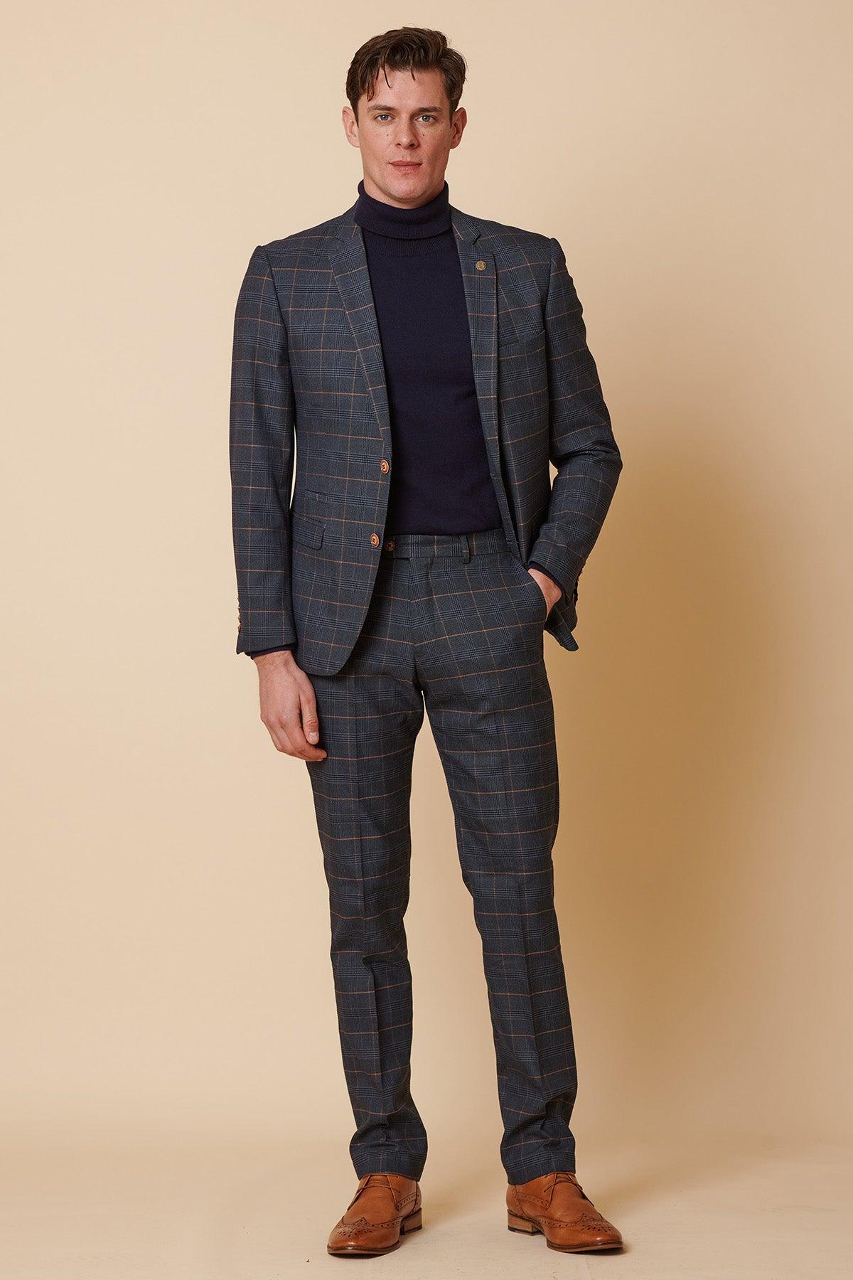 A man stands against a beige background wearing the Marc Darcy JENSON - Marine Navy Check Two Piece Suit, paired with a navy turtleneck and brown dress shoes. One hand is in his pocket as he looks directly at the camera.