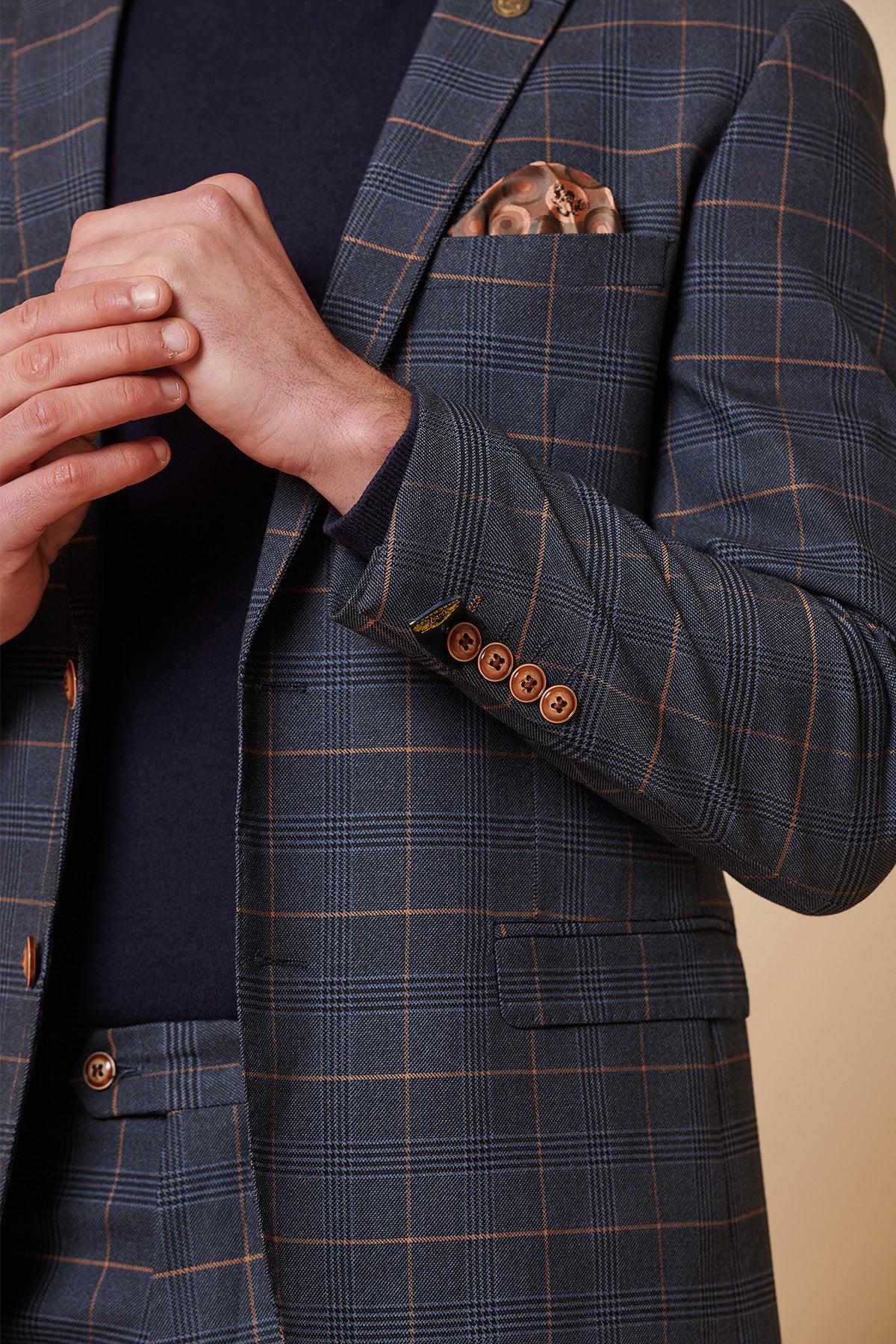 A person wearing the Marc Darcy JENSON Marine Navy Check Two Piece Suit stands against a neutral background, adjusting their jacket sleeve. The suit features brown buttons and a matching pocket square, paired with a dark shirt.