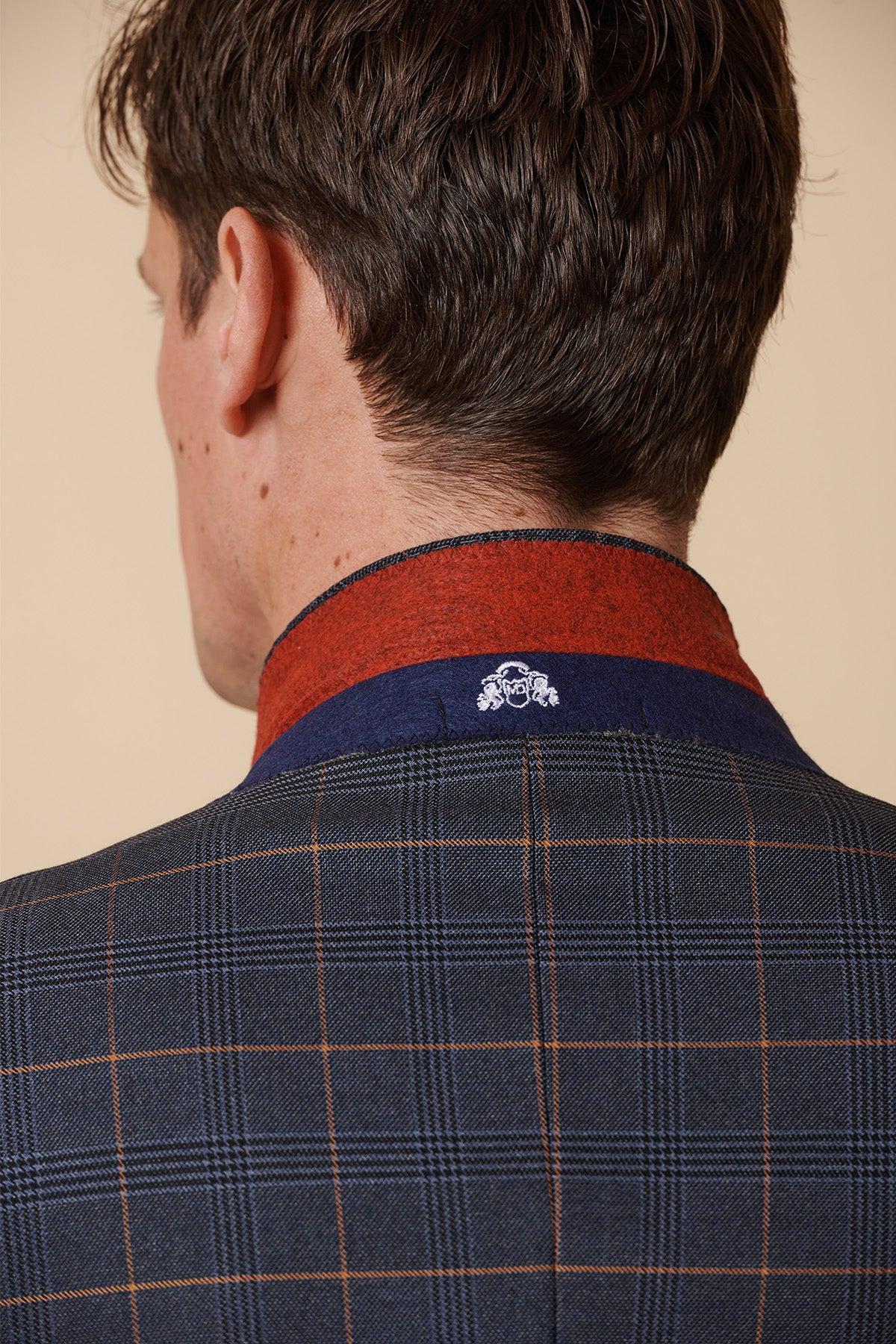 A man seen from behind wears the Marc Darcy JENSON Marine Navy Check Two Piece Suit with a dark plaid blazer and red-blue collar, accented by a white embroidered crest. The beige background contrasts with the suit's sharp style.