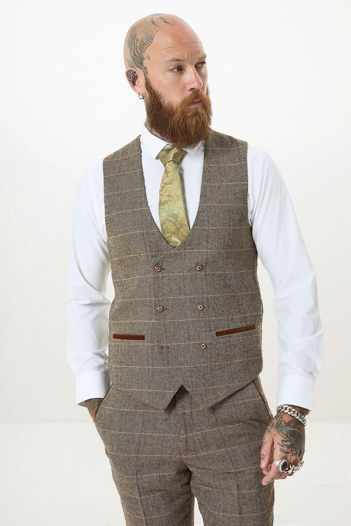 A bearded man with tattoos and piercings models the marcdarcy TED Tan Tweed Check Double Breasted Waistcoat with matching trousers, a white shirt, and patterned green tie, standing against a plain white background and looking to the side.