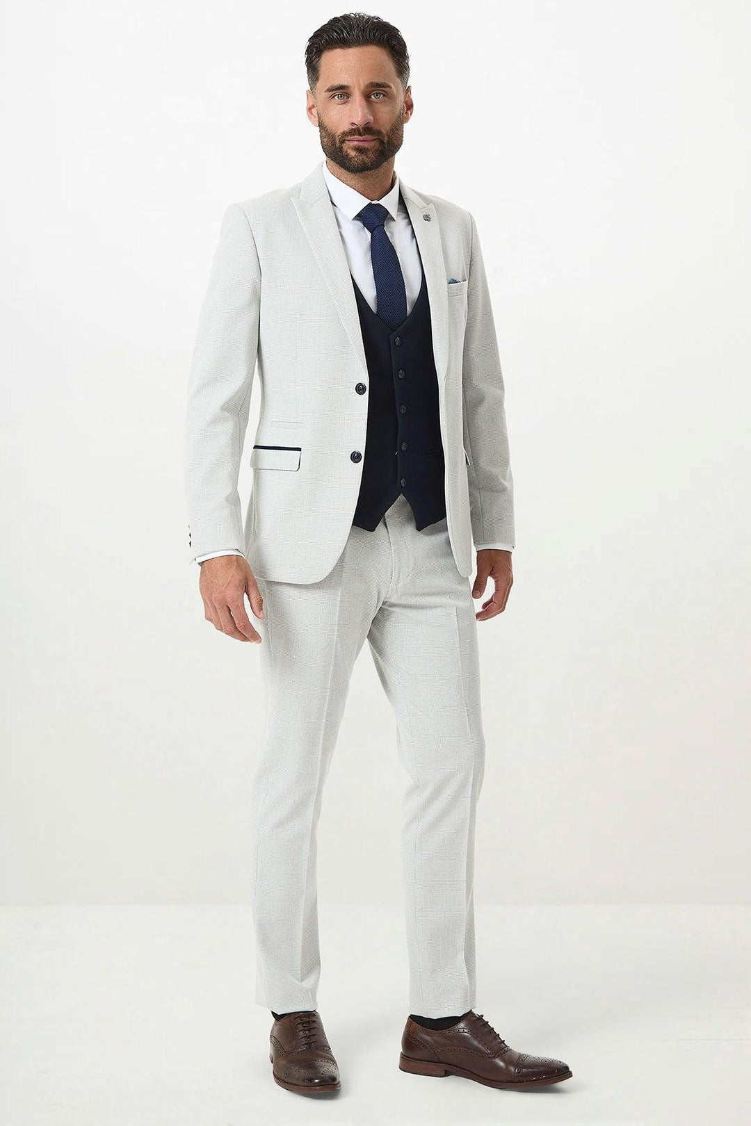 A man stands against a white background wearing the Marc Darcy BROMLEY Stone Check Suit with a Kelvin Navy Waistcoat, white shirt, navy blue tie, and brown dress shoes.