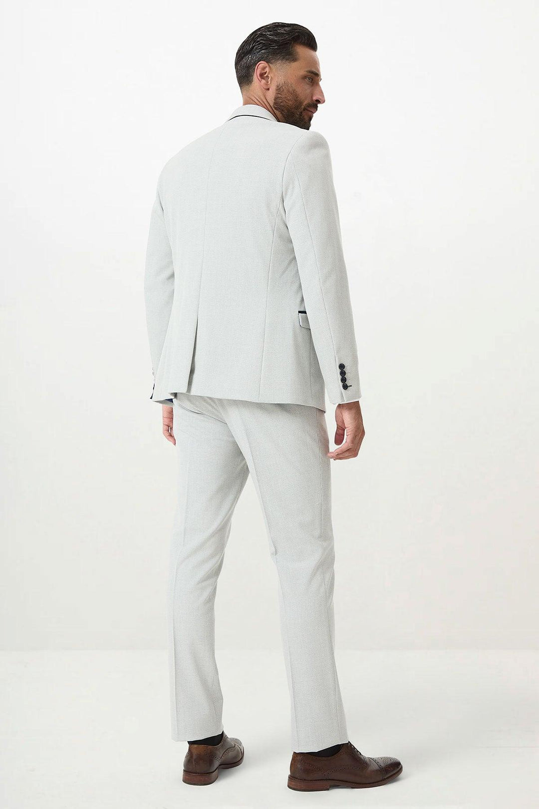 A man with dark hair and a beard models the Marc Darcy BROMLEY - Stone Check Suit with Kelvin Navy Waistcoat, styled in a slim fit with brown dress shoes, standing with his back to the camera against a plain white background.