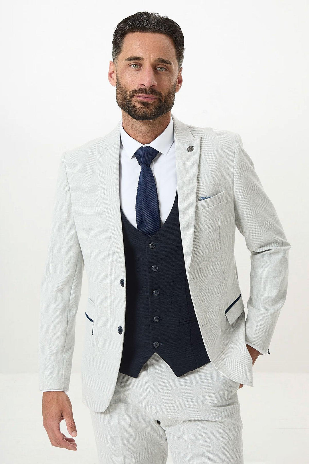 A man with dark hair and a beard wears the Marc Darcy BROMLEY Stone Check Suit paired with a Kelvin Navy Waistcoat, white shirt, and navy tie. He stands against a plain white background, looking confidently at the camera.