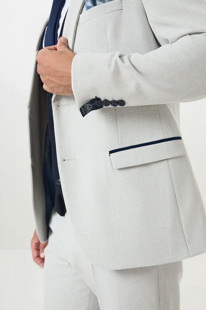 A person wearing the Marc Darcy BROMLEY Stone Check Suit with a Kelvin Navy Waistcoat, blue tie, and patterned pocket square, holding the jacket front. Focus is on the suits details and upper body.
