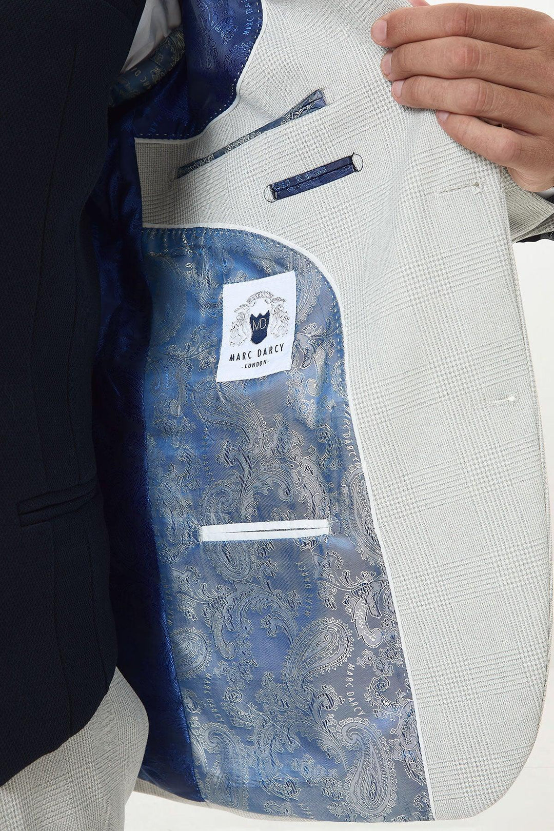 A person holds open a BROMLEY - Stone Check Suit jacket by Marc Darcy, revealing a blue paisley lining and a Marc Darcy label stitched inside the inner pocket.
