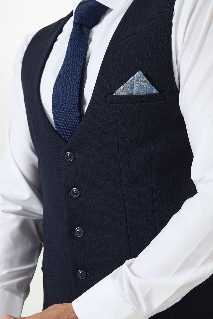 A man wears a Marc Darcy BROMLEY Stone Check Suit with a Kelvin Navy Waistcoat, white shirt, textured navy tie, and blue patterned pocket square, shown from shoulders to waist.
