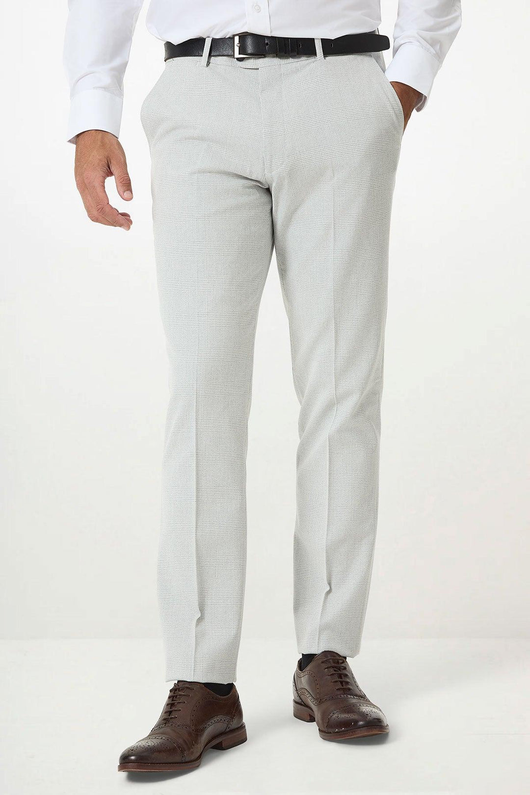 A man dressed in the Marc Darcy BROMLEY Stone Check Suit with Kelvin Navy Waistcoat stands against a plain background, wearing a white shirt, black belt, and brown dress shoes, one hand in his pocket.