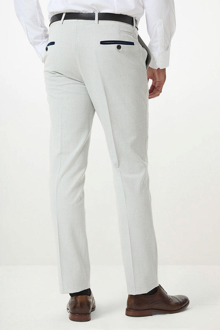 A man wearing Marc Darcys BROMLEY Stone Check Suit pants and a white shirt stands facing away; the light gray checked trousers feature black trim on the back pockets.