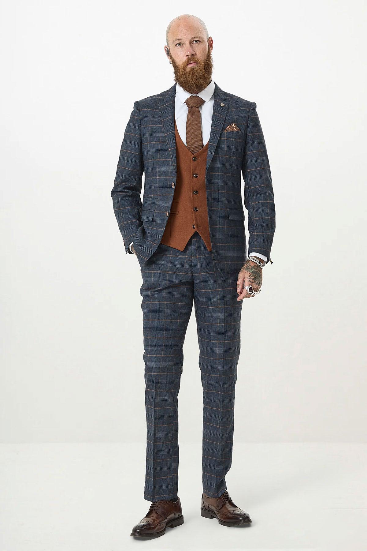 A bearded man models the marcdarcy JENSON - Marine Navy Check Suit with a Kelvin Tan Waistcoat, brown tie, and brown dress shoes, standing against a plain white background with one hand in his pocket and the other at his side.