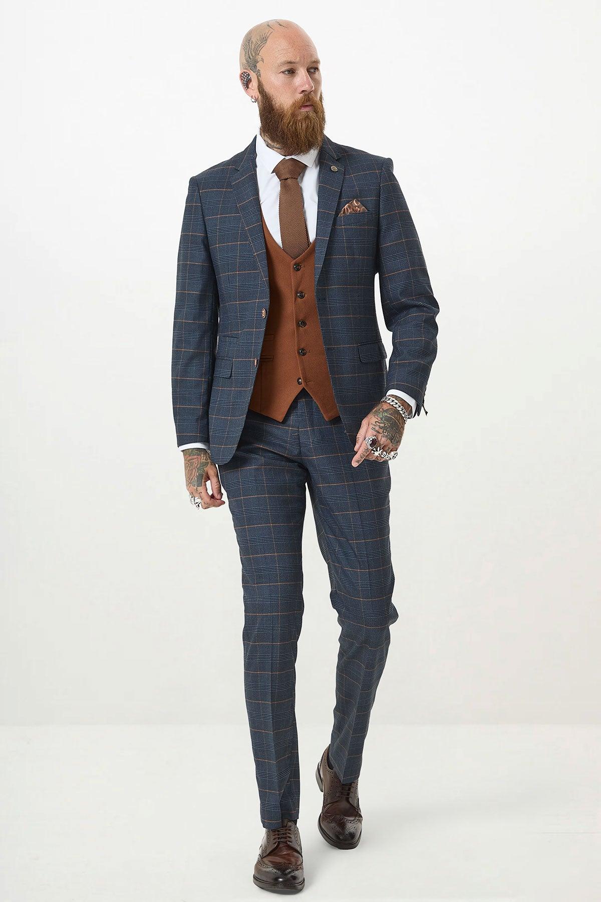 A bearded, tattooed man wears the marcdarcy JENSON Marine Navy Check Suit with a Kelvin Tan Waistcoat, white shirt, brown tie, and brown dress shoes as he walks confidently against a plain white background.