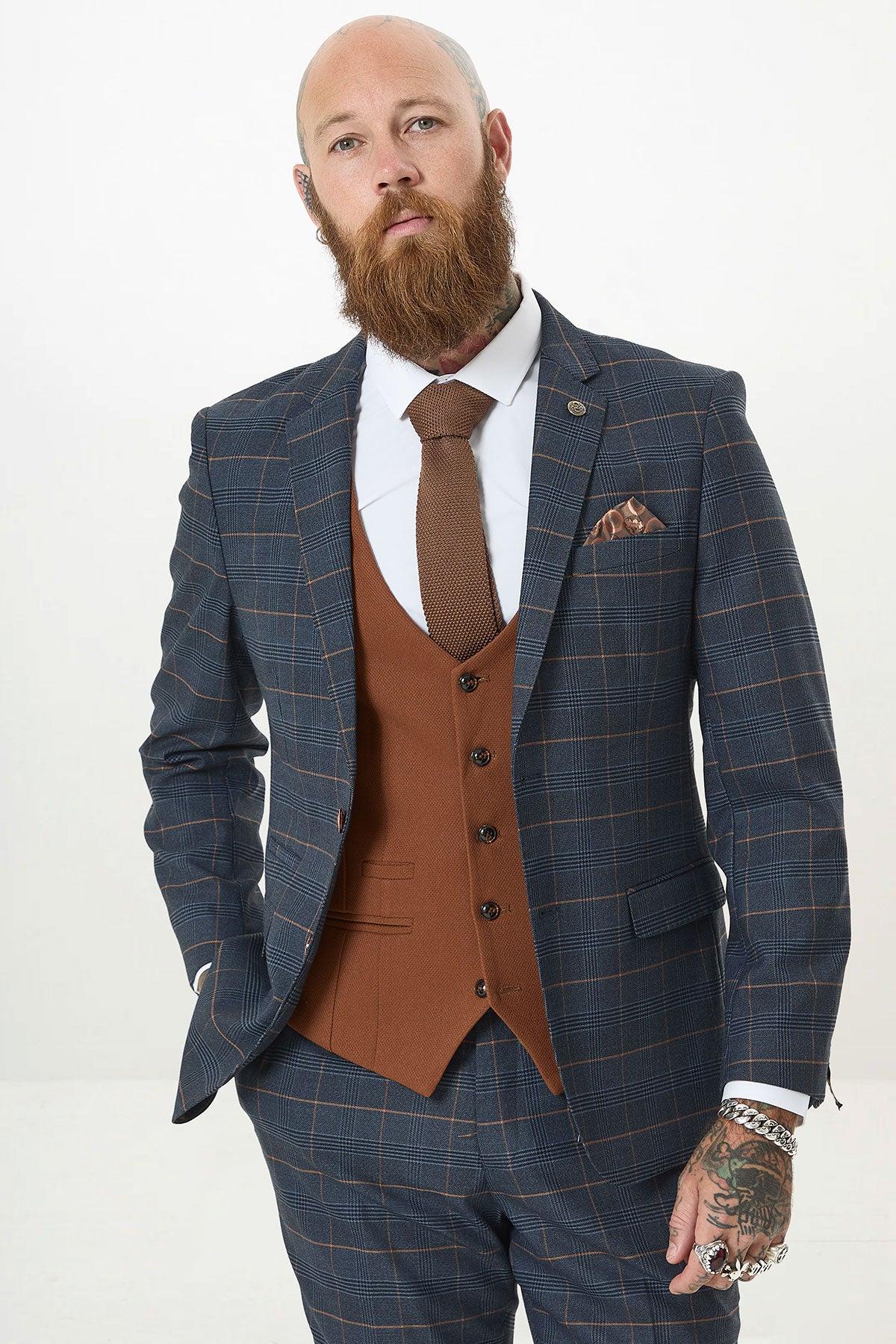 A bearded man with tattoos wears the marcdarcy JENSON - Marine Navy Check Suit with Kelvin Tan Waistcoat, brown tie, and white shirt, standing with one hand in his pocket against a plain white background.