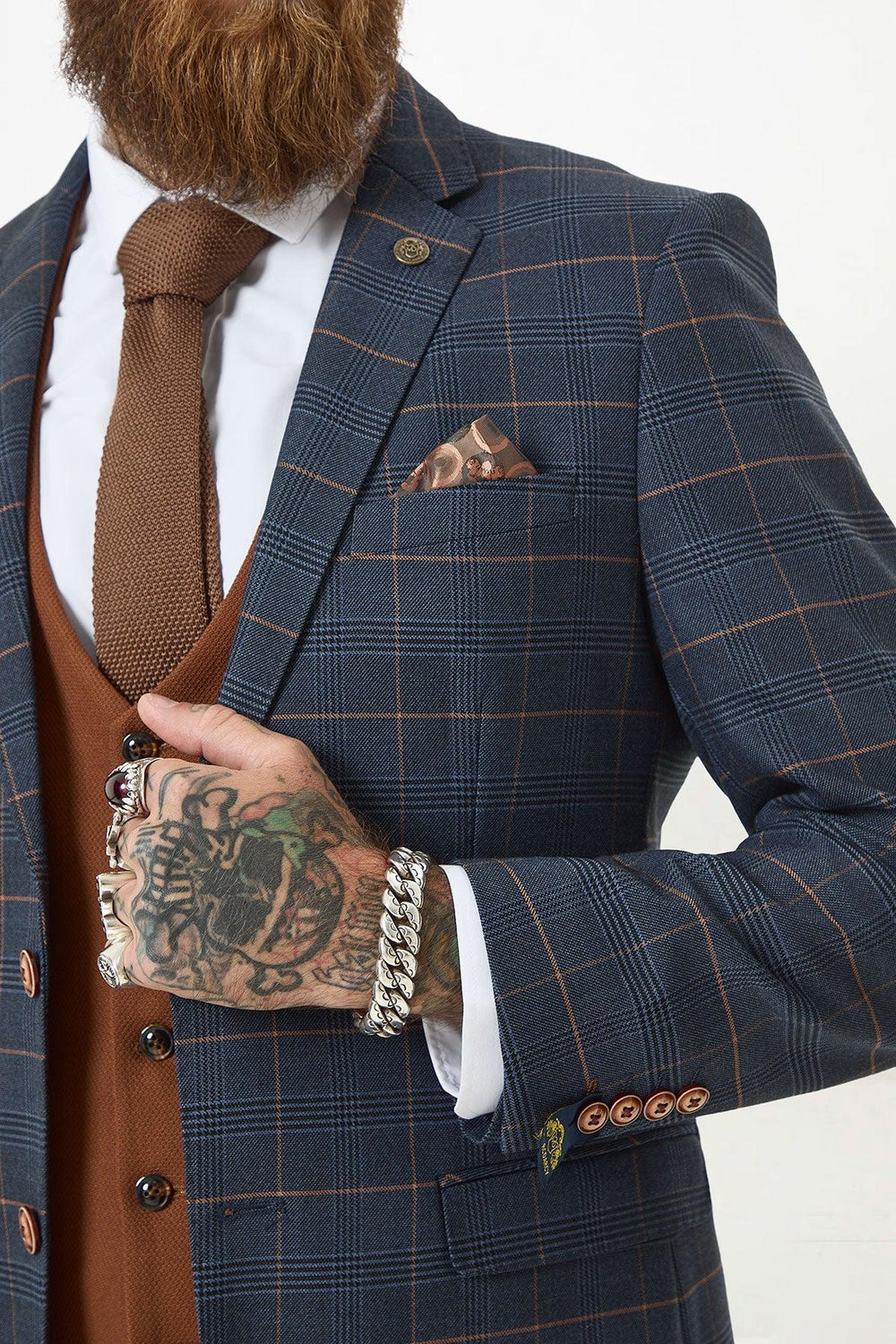 A man wears the marcdarcy JENSON Marine Navy Check Suit with Kelvin Tan Waistcoat, brown tie, and pocket square. He sports a beard, tattooed hand, silver rings and bracelets.
