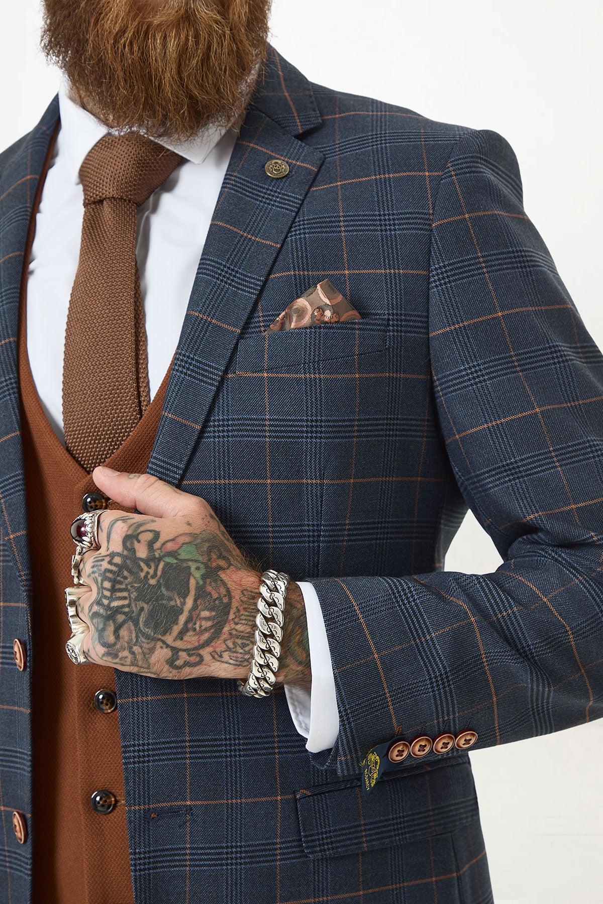 A man in the marcdarcy JENSON Marine Navy Check Suit with a Kelvin Tan Waistcoat, styled with a white shirt, brown tie, brown beard, visible hand tattoos, silver bracelets and rings, and a patterned pocket square.