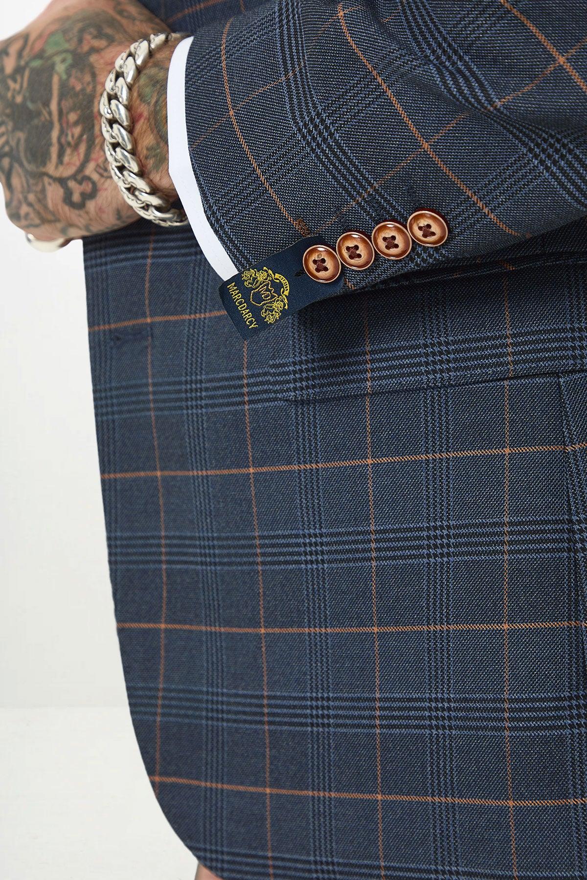 A close-up of a person wearing the marcdarcy JENSON Marine Navy Check Suit with Kelvin Tan Waistcoat, featuring brown cuff buttons, a visible logo tag, orange check lines, and a silver chain bracelet on a tattooed wrist.