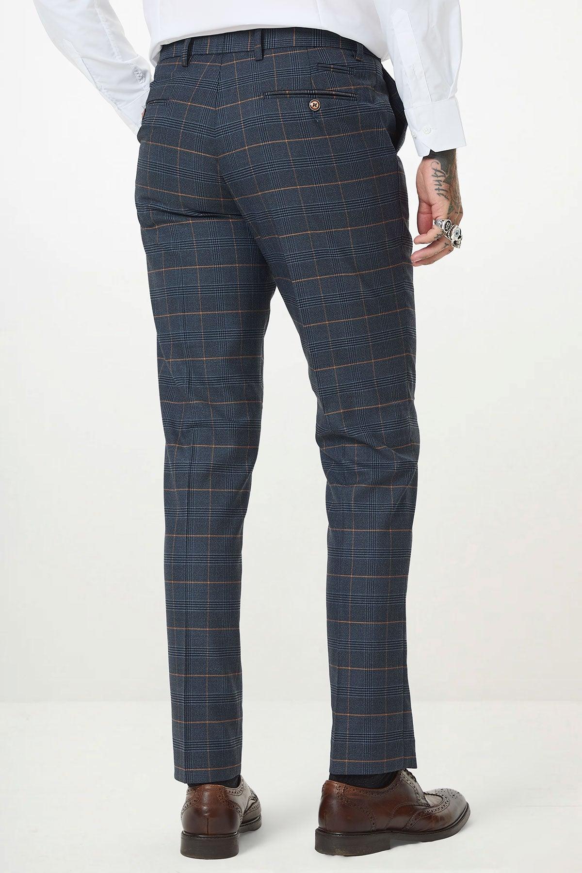 A person wearing marcdarcy’s JENSON - Marine Navy Check Suit With Kelvin Tan Waistcoat, a white shirt, and brown dress shoes stands facing away from the camera. The suit features a subtle check pattern and back pockets, with tattoos on their left hand.
