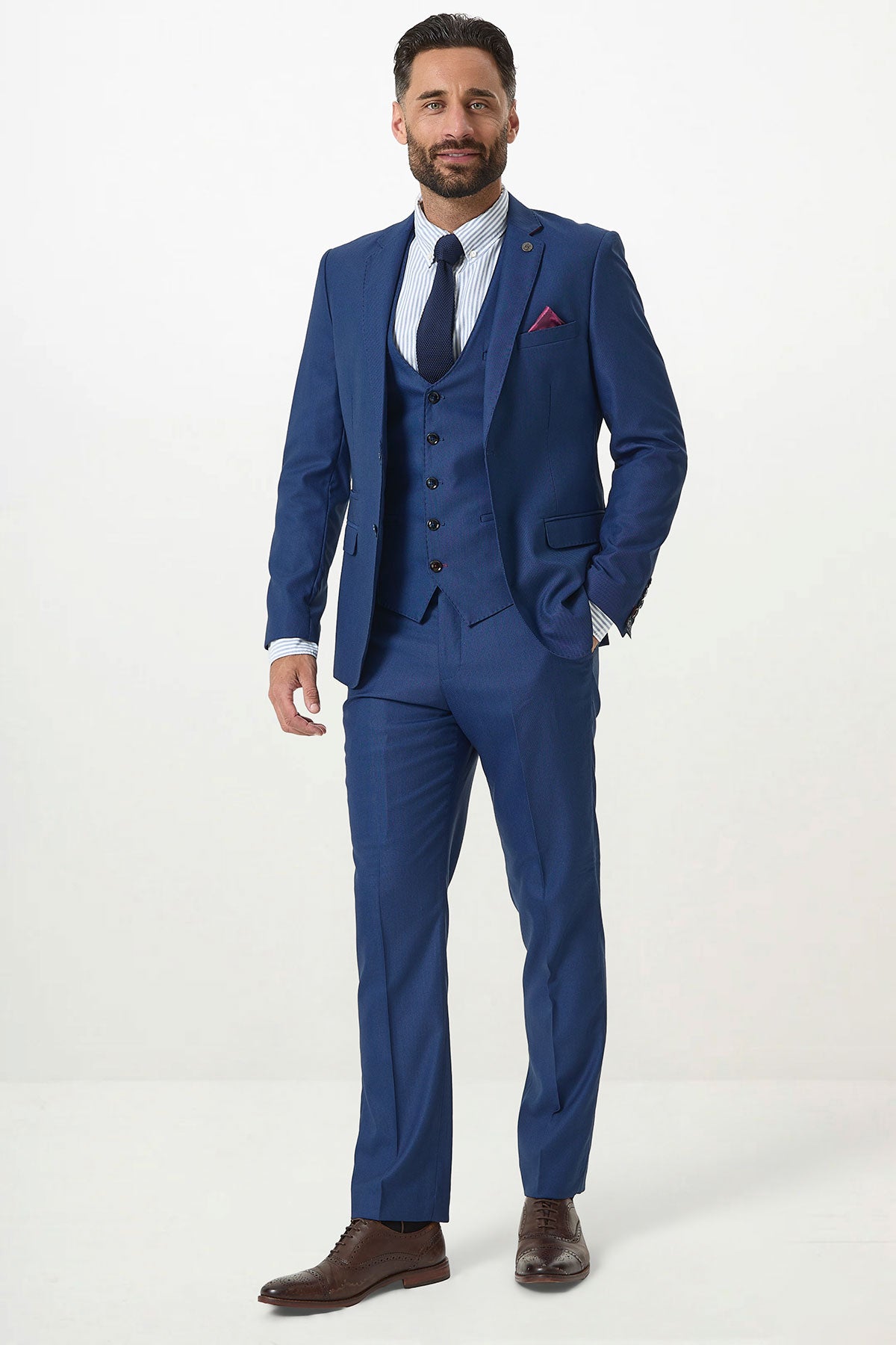 A man wears the Marc Darcy DANNY Royal Blue Three Piece Suit with a single-breasted waistcoat, striped shirt, navy tie, and brown shoes. He stands against a white background, smiling—a stylish modern update for any workwear wardrobe.