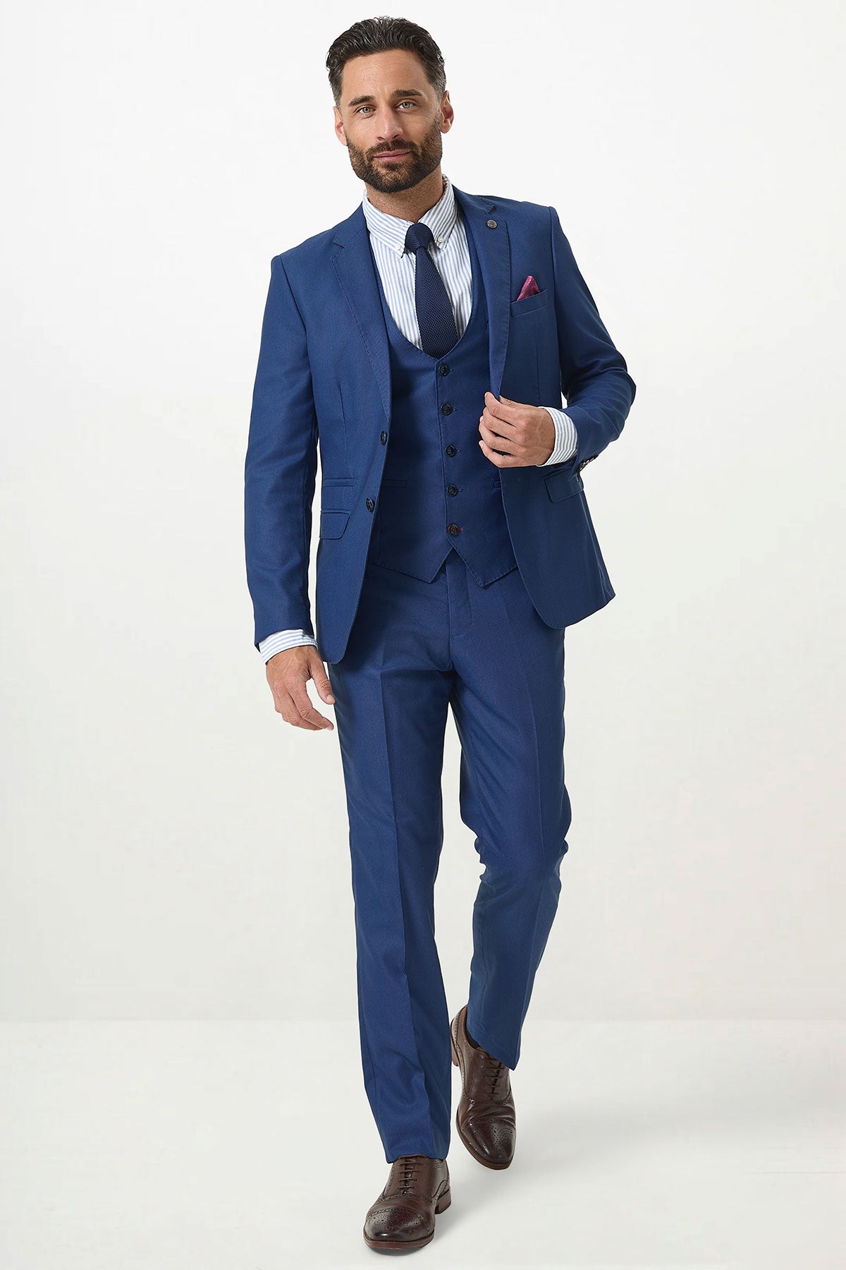 A man with a trimmed beard stands against a white background, wearing the Marc Darcy DANNY Royal Blue Three Piece Suit with single breasted waistcoat, paired with a striped shirt, navy tie, and brown dress shoes—showcasing sharp modern workwear style.