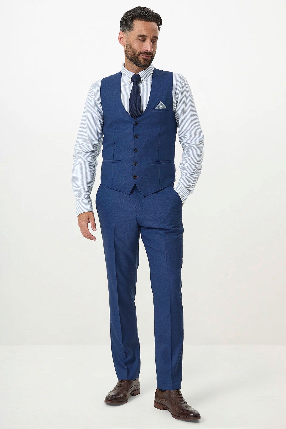 A man wears the Marc Darcy DANNY Royal Blue Single Breasted Waistcoat with a striped shirt, navy tie, and brown dress shoes, standing against a white background with one hand in his pocket.