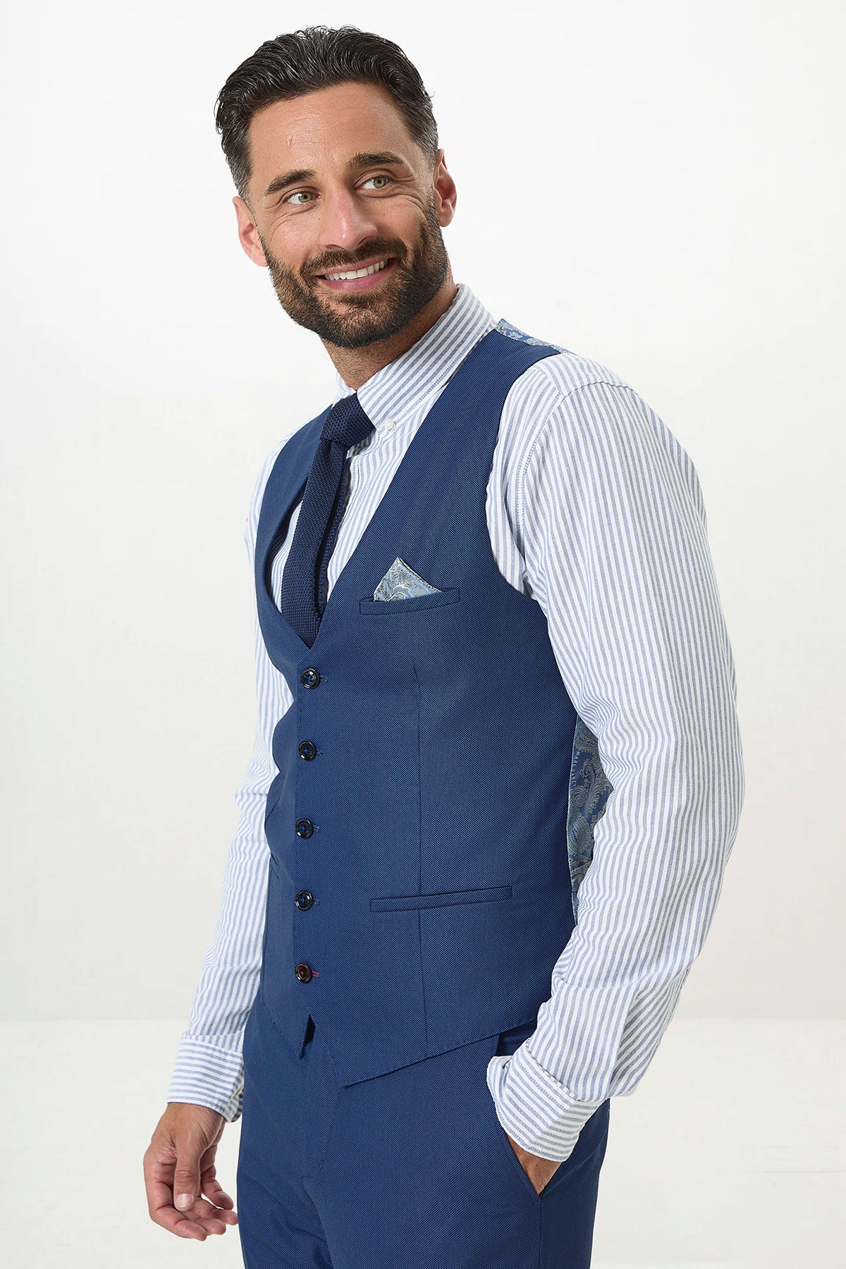 A man with dark hair and a beard wears the Marc Darcy DANNY Royal Blue Three Piece Suit with a single breasted waistcoat, complemented by a blue and white striped shirt, tie, and pocket square. A sharp choice for any workwear wardrobe.