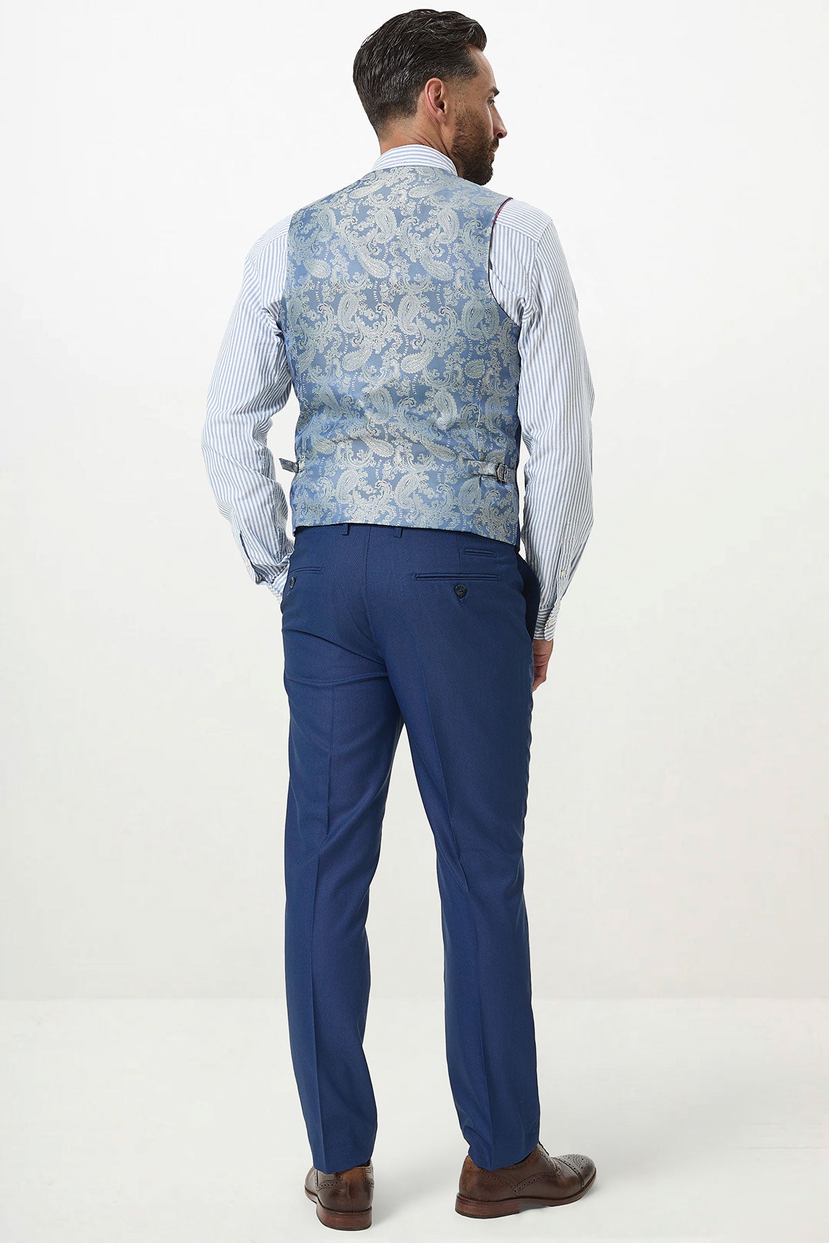 A man wears the Marc Darcy DANNY Royal Blue Single Breasted Waistcoat over a striped shirt with blue trousers and brown shoes, standing against a plain white background.
