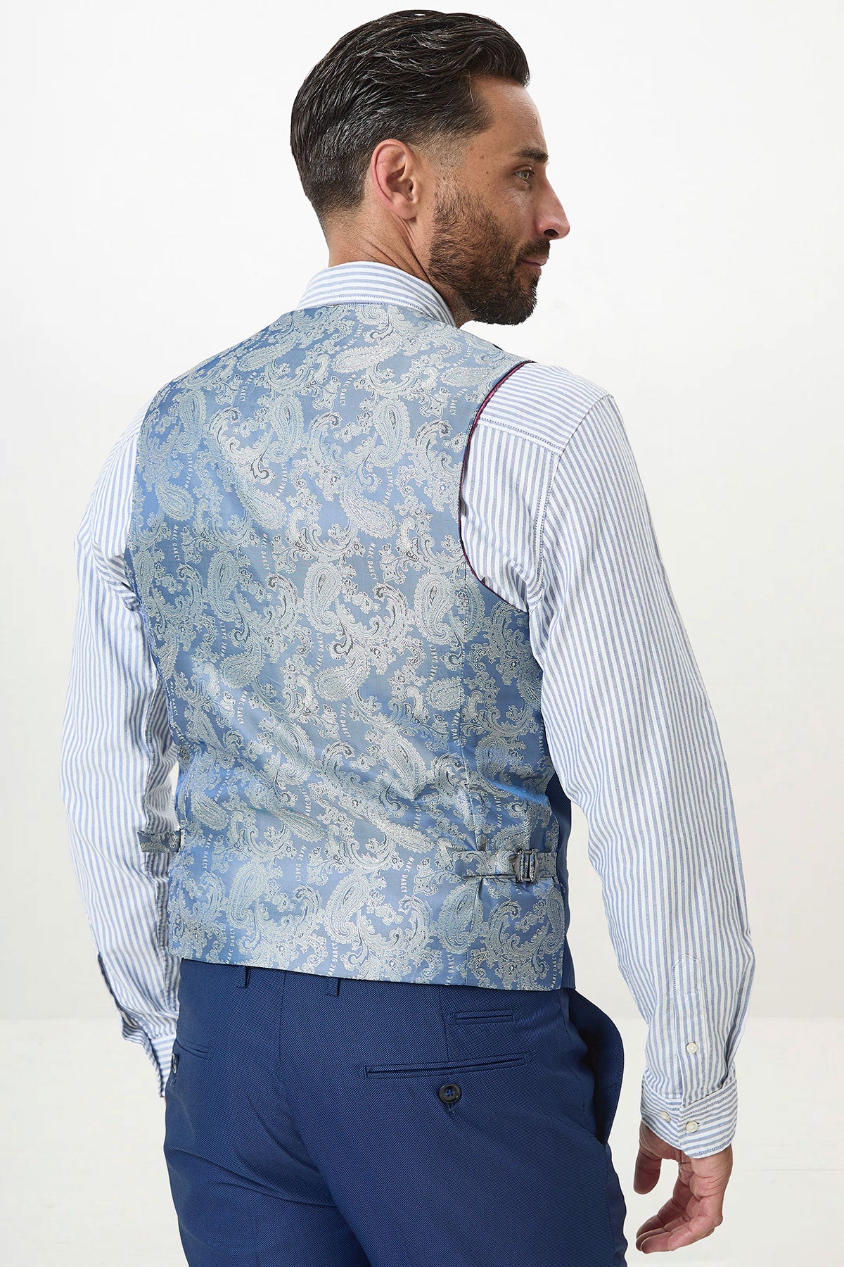 A man with dark hair and a beard is seen from behind wearing the Marc Darcy DANNY Royal Blue Single Breasted Waistcoat over a blue striped dress shirt and blue trousers, standing against a plain white background.