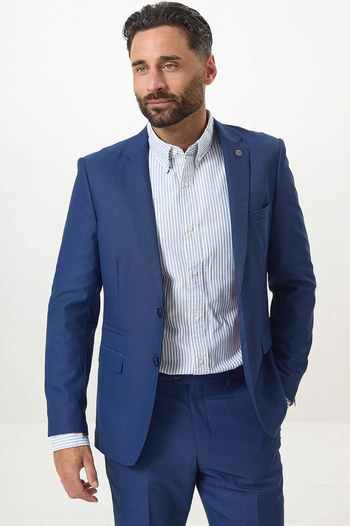 A man with dark hair and a beard wears the Marc Darcy DANNY Royal Blue Two Piece Suit with a light blue and white striped shirt. He stands against a plain, light background, looking slightly to the side with one hand in his pocket.